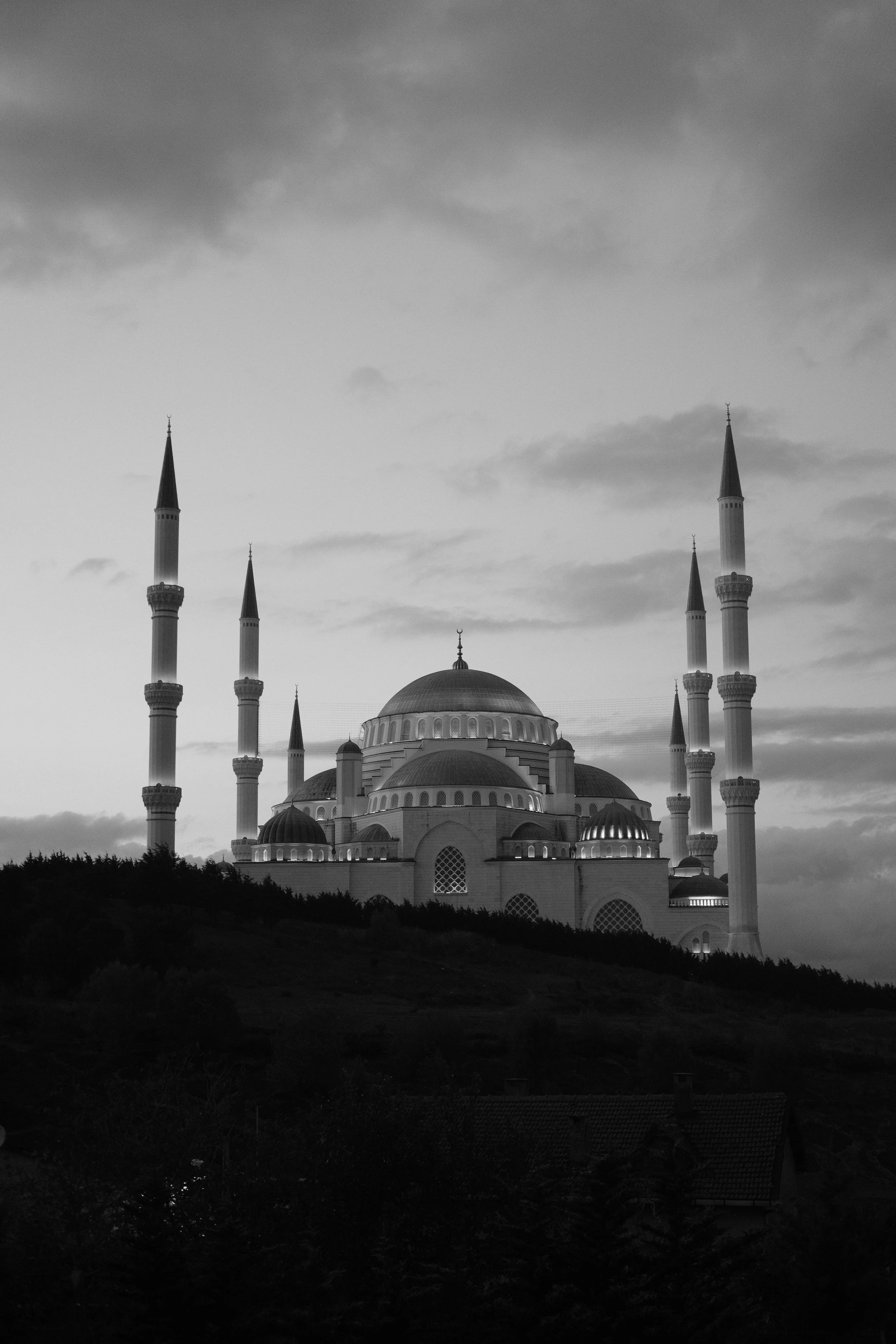 Schwarz-Weiß-Bild einer Moschee mit großen Kuppeln und sechs Minaretten, auf einem Hügel mit Bäumen und einem Haus im Vordergrund.