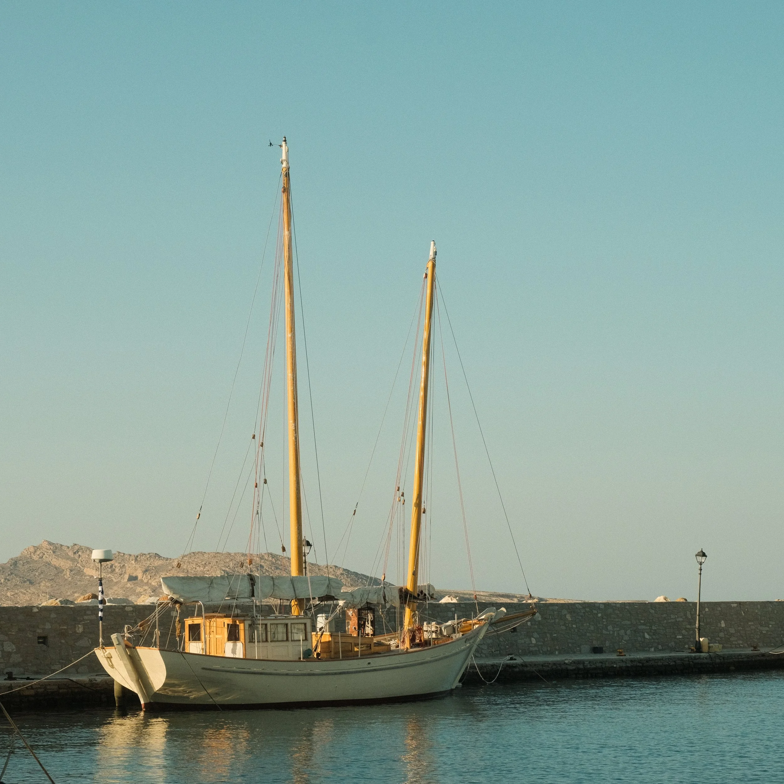Ein Segelboot liegt am Kai neben einer Mauer mit Straßenlaternen, im Hintergrund Berge, bei ruhigem Wasser und klarem Himmel.