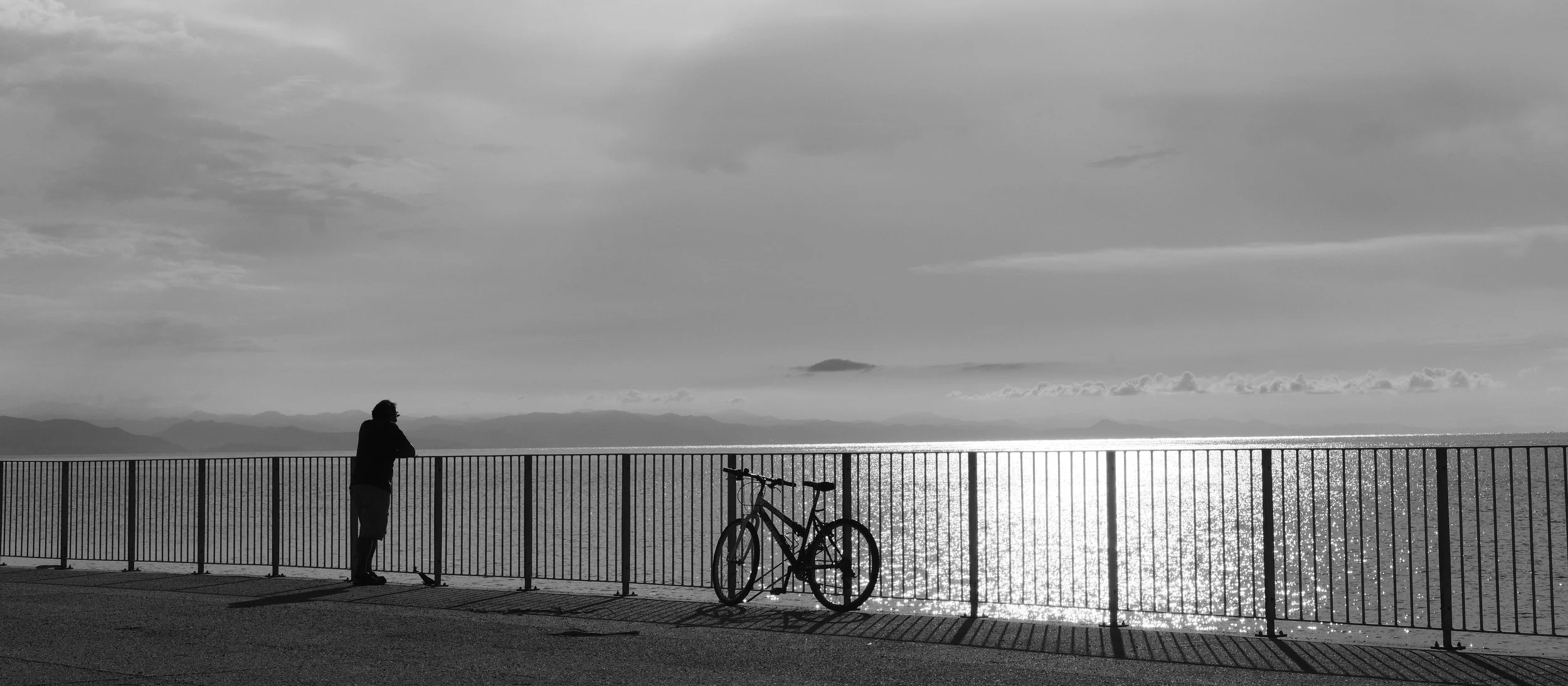 Eine schwarze Silhouette einer Person, die an einer Straßengrenze mit Blick auf den Ozean steht, neben einem Fahrrad. Der Himmel ist bewölkt und das Wasser reflektiert das Licht.