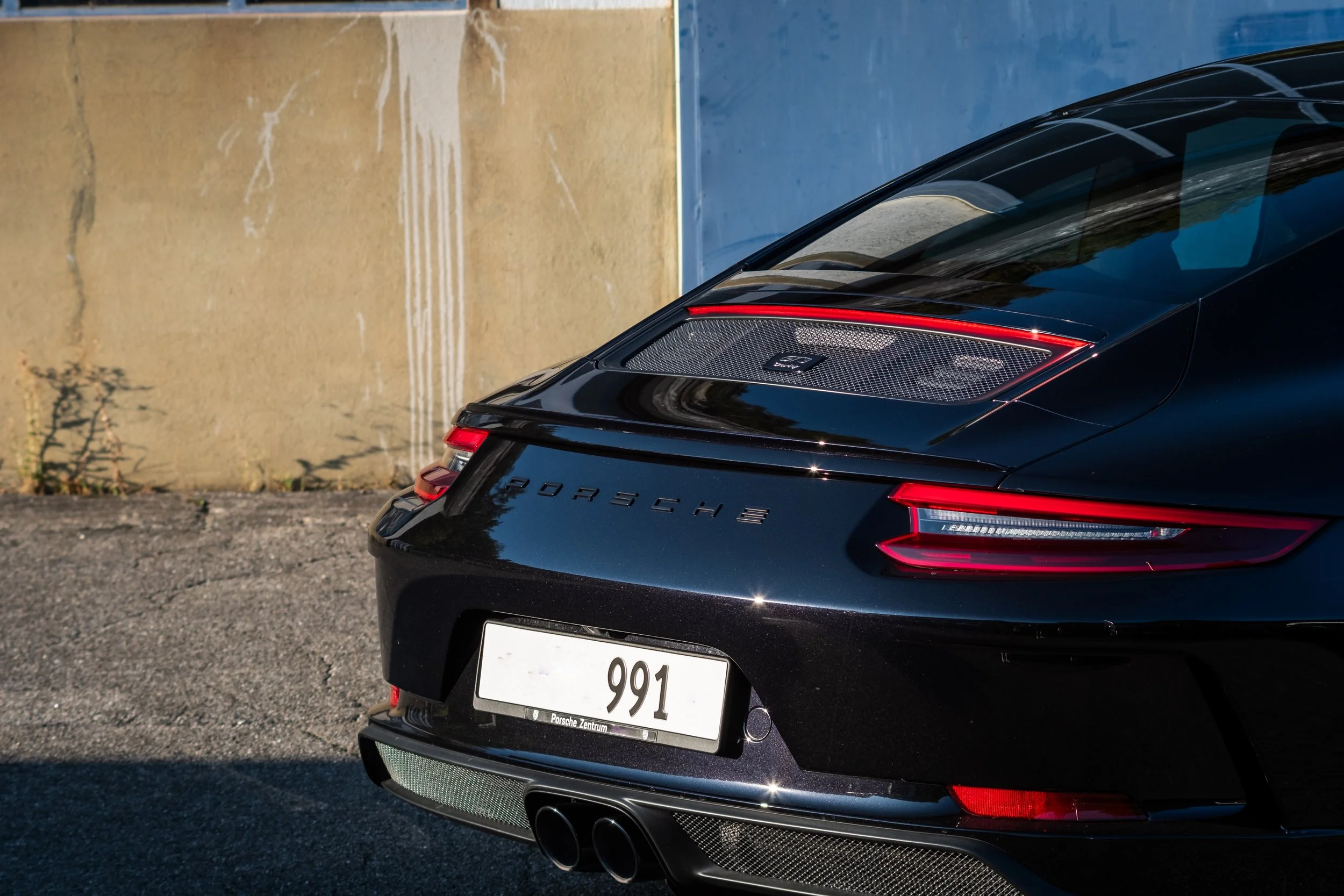 Rückansicht eines schwarzen Porsche 911 mit deutschem Kennzeichen, auf einem Asphaltplatz vor einer Mauer.