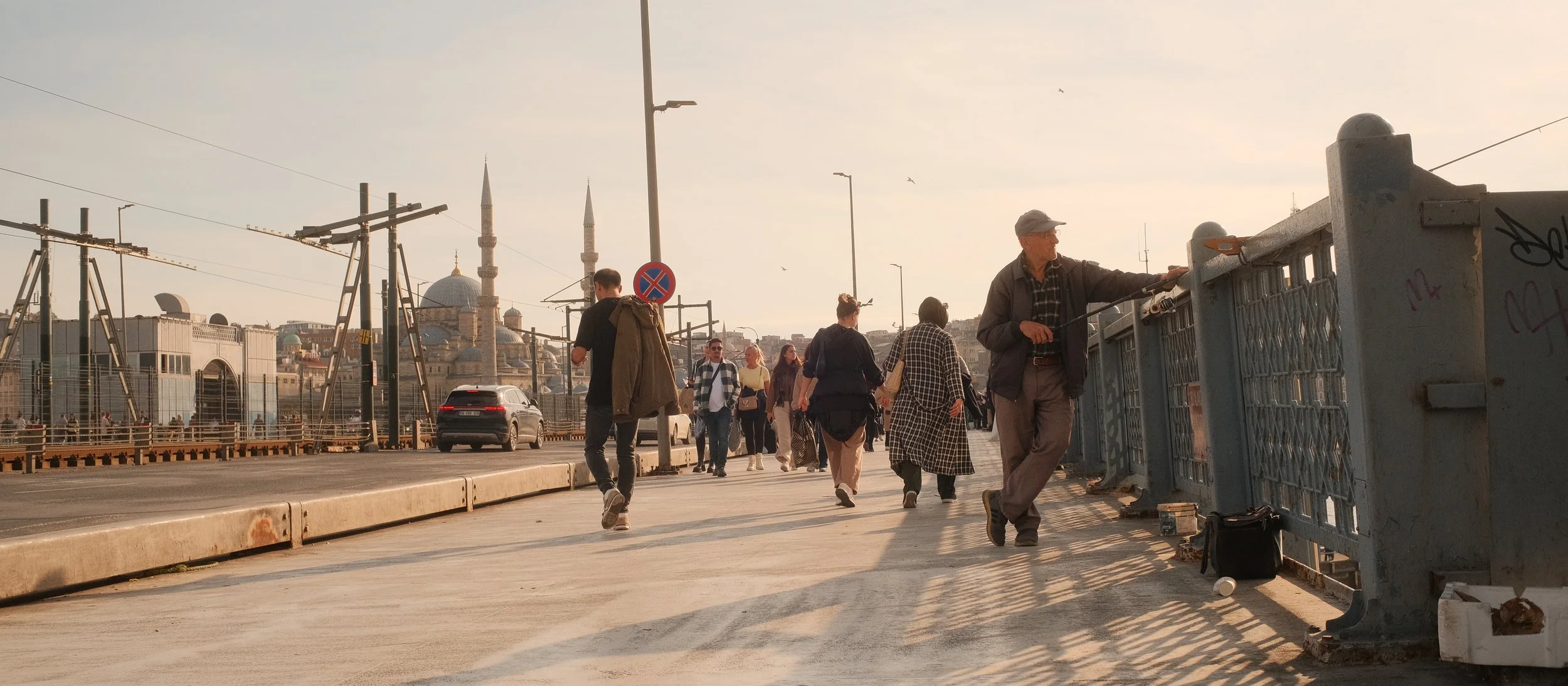 Menschen gehen auf einer Brücke, einige schauen zum Fluss, im Hintergrund sind Moscheen mit Minaretten sichtbar, das Sonnenlicht wirft lange Schatten.