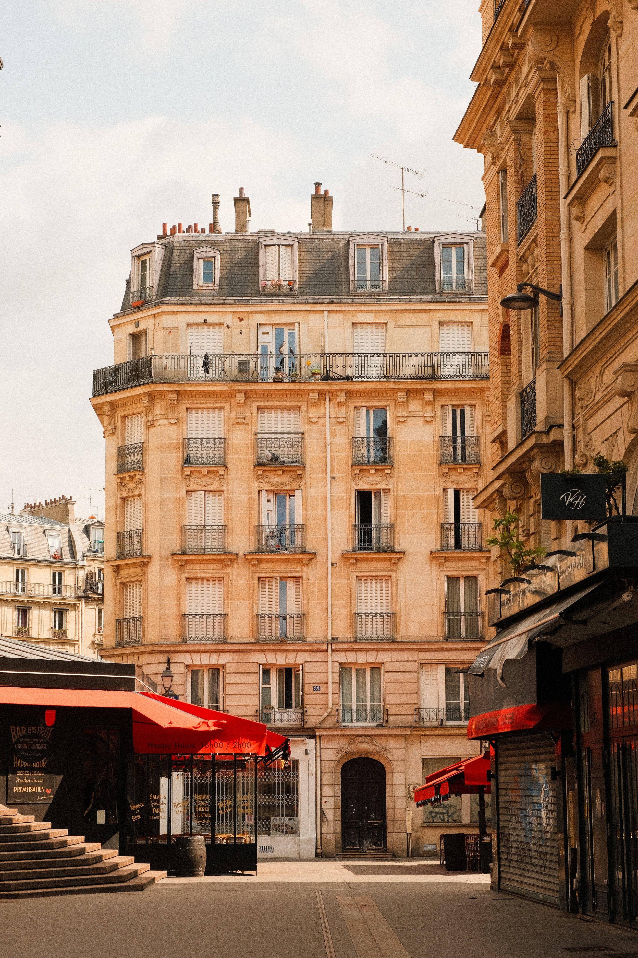 Ein Parisianisches Gebäude mit mehreren Stockwerken, Fensterläden und Balkonverzierungen, umgeben von Geschäften mit roten Markisen
