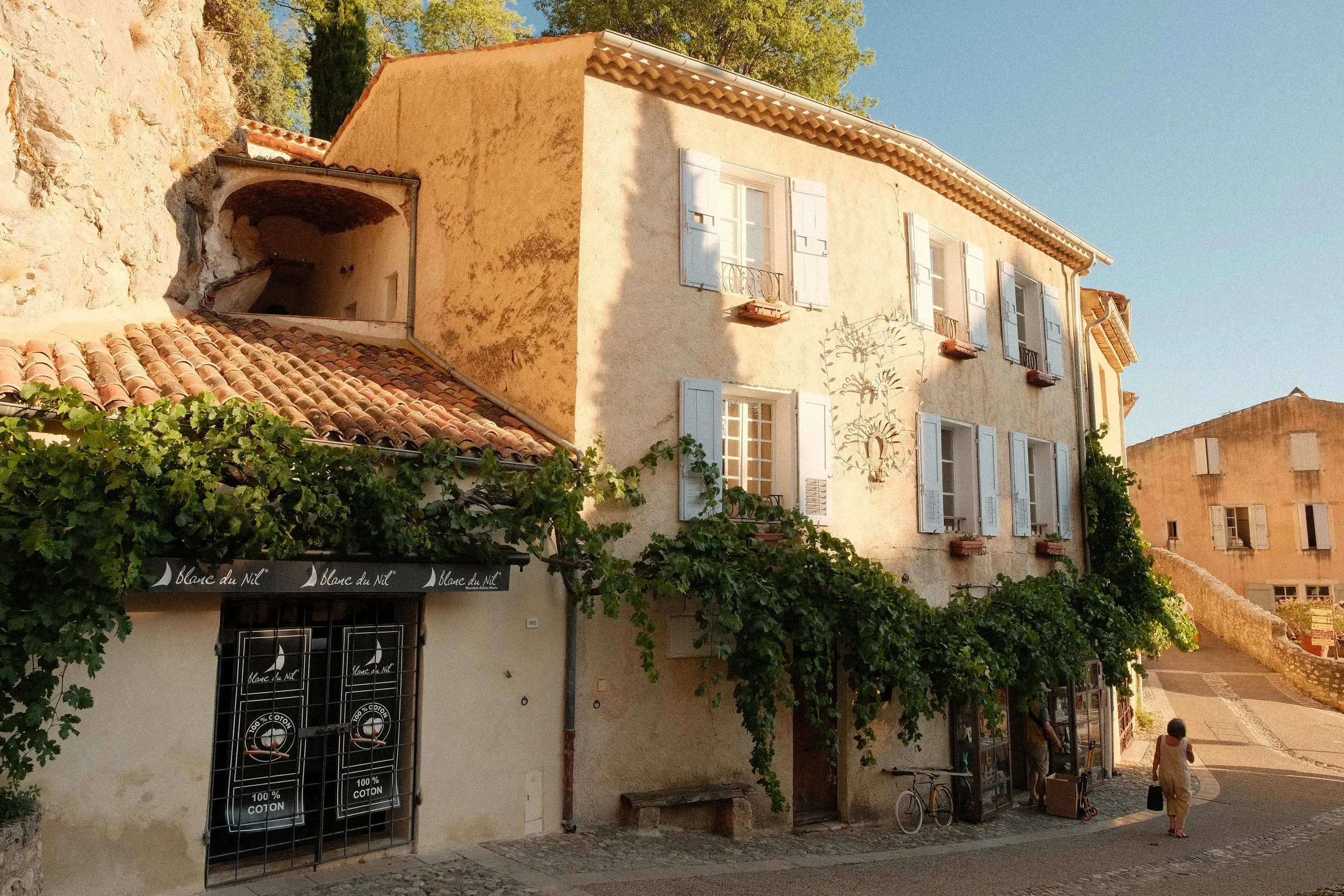 Ein altes Gebäude mit weißen Fensterläden, umgeben von Grün, in einer sonnigen kleinen Stadt. Es gibt Leute, die die Straße entlanggehen, und einen Fahrrad am Gebäude.