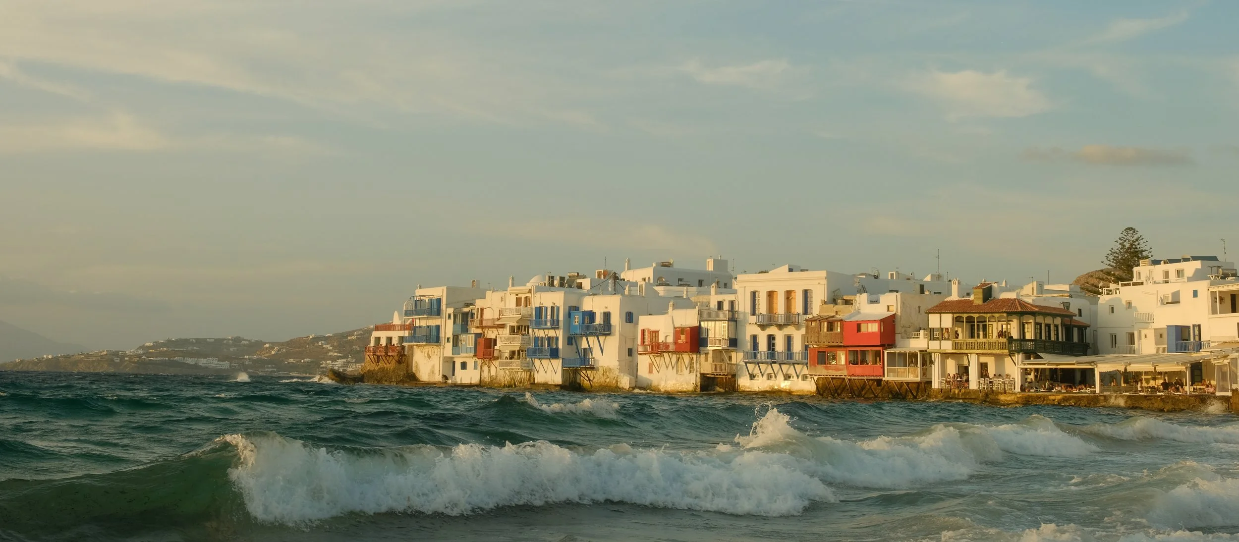 Blick auf eine weiße Küstenstadt mit bunten Gebäuden, die direkt am Wasser liegen, bei Sonnenuntergang, mit Wellen im Vordergrund.