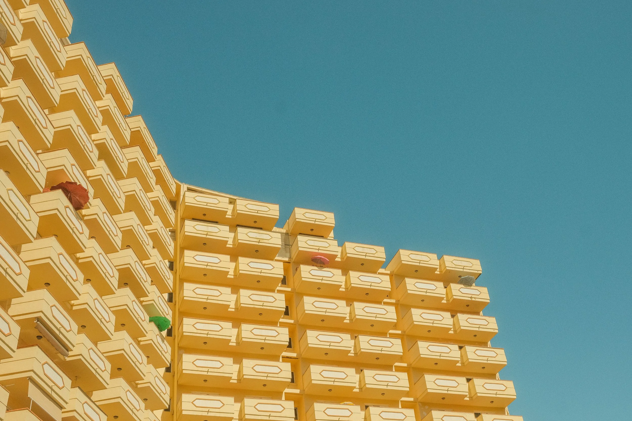 Blick auf die Ecke eines modernen, gelben Hochhauses mit mehreren Balkonen gegen blauen Himmel.