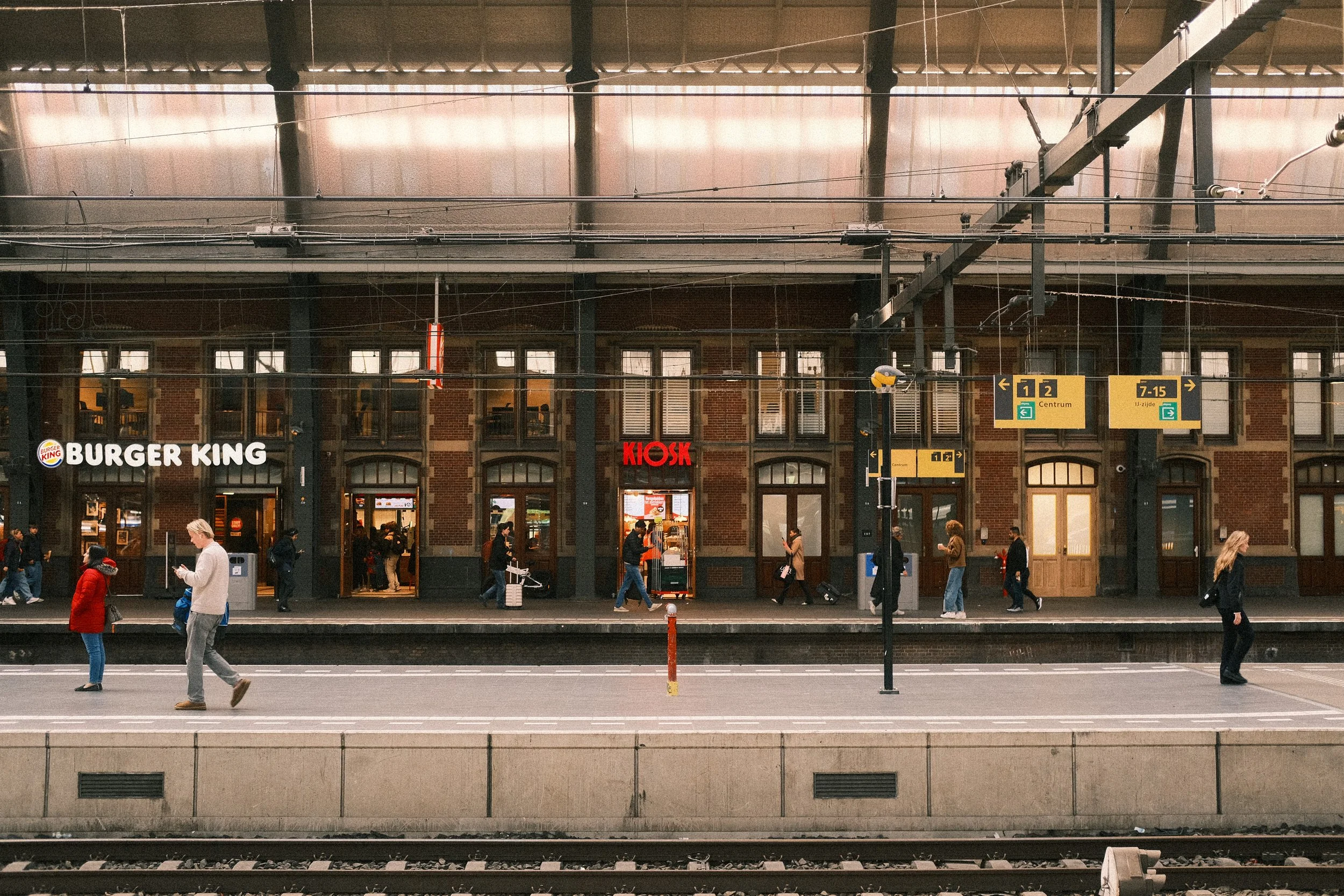 Menschen warten am Bahnsteig vor einem alten Backsteinbahnhof mit Signalen und einem Burger King und Kiosk im Hintergrund.