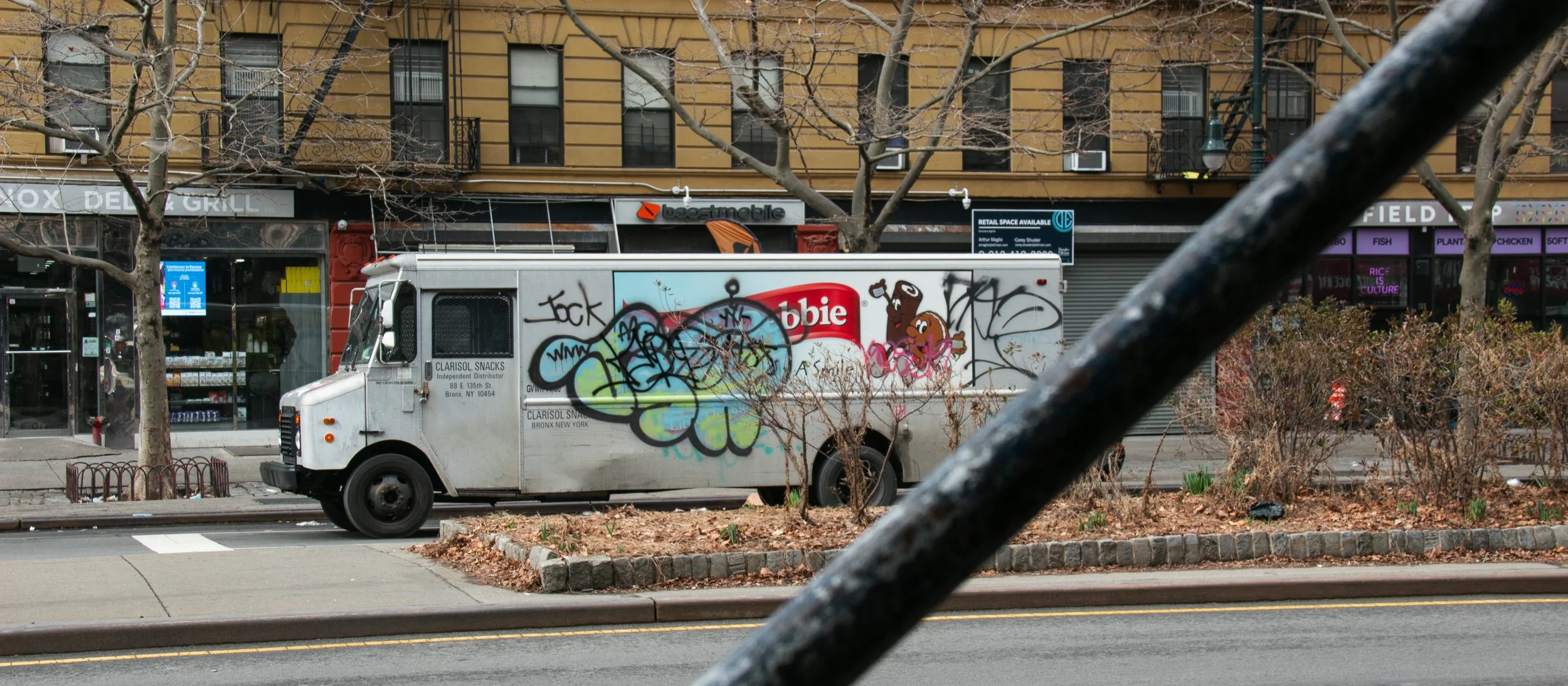 Ein Grauer Lieferwagen ist auf einer Stadtstraße geparkt, die mit grauen Gehwegen und einer kleinen Baumschutzanlage versehen ist. Der Lieferwagen ist mit Graffiti bemalt, darunter ein lachender Brauster mit Kochmütze, und hat ein Logo von 'Clarisol 