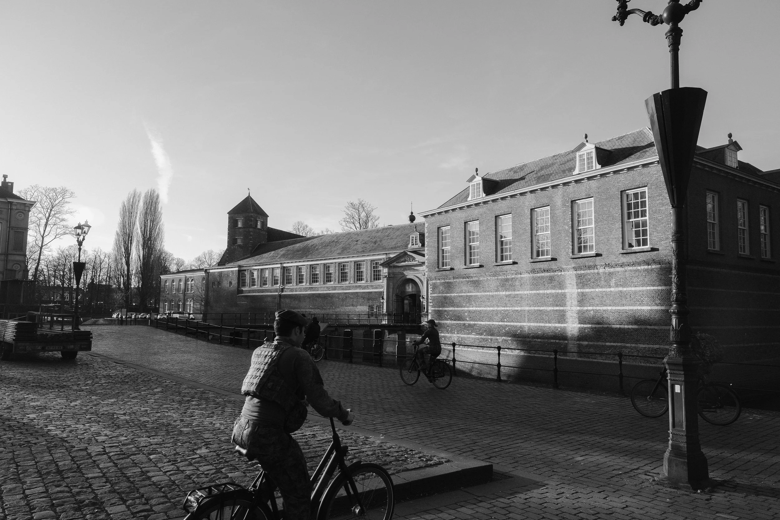 Blick auf eine historische Gebäude mit Zinnen und einem runden Turm, Fahrradfahrer und Fußgänger auf Kopfsteinpflasterstraße, Laterne im Vordergrund, in schwarz-weiß aufgenommen.