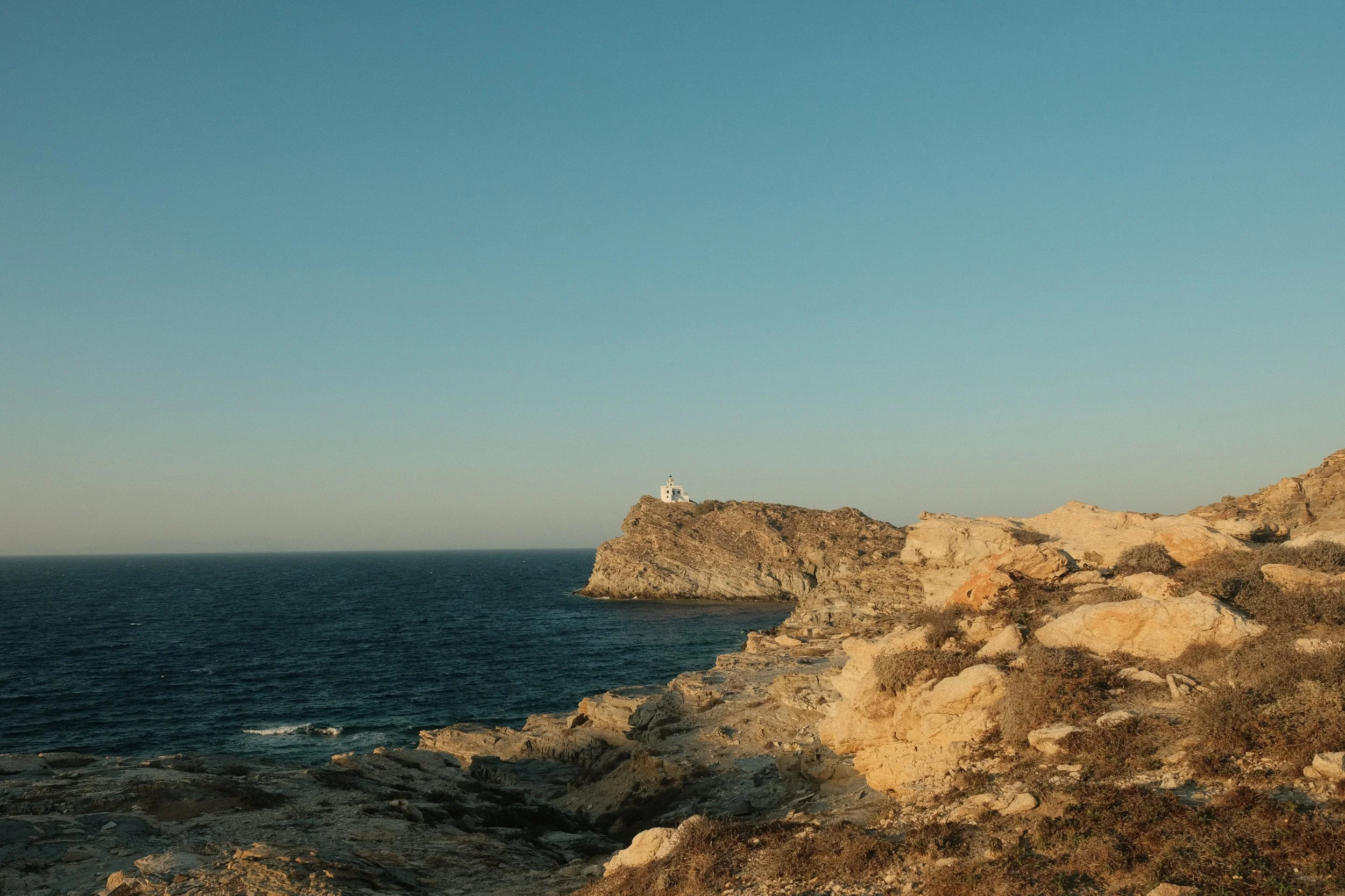 Ein Küstenabschnitt mit Felsen und einem kleinen weißen Leuchtturm auf einer Klippe im Meer bei Sonnenuntergang.