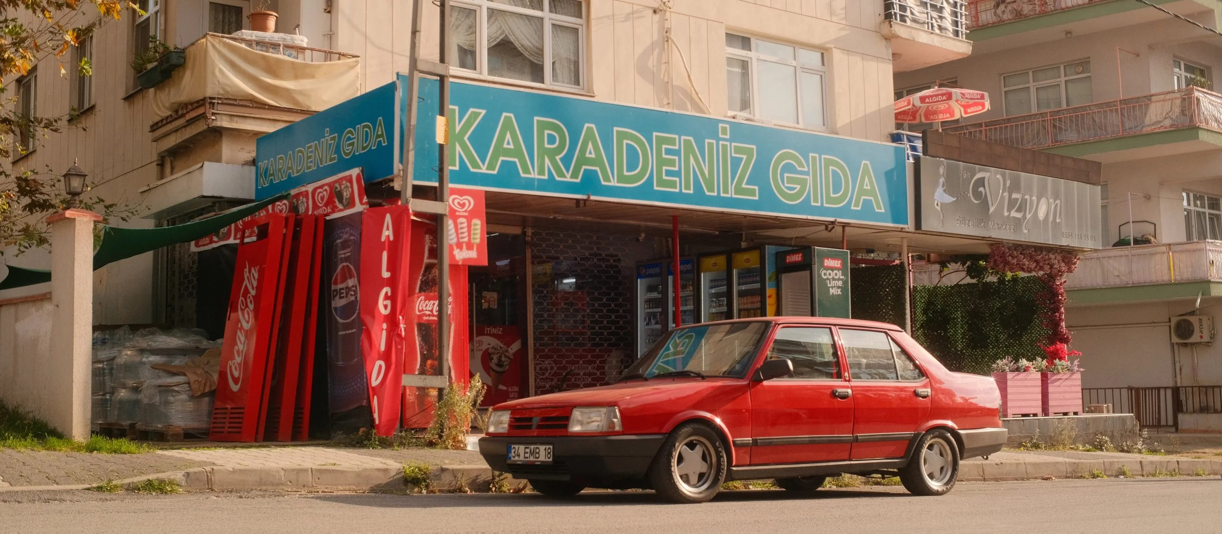 Ein rotes Auto parkt vor einem Lebensmittelgeschäft mit einem großen blauen Schild, das 'KARADENİZ GIDA' sagt. Das Geschäft befindet sich in einem Mehrfamilienhaus, mit Balkon und anderen Geschäften im Hintergrund.