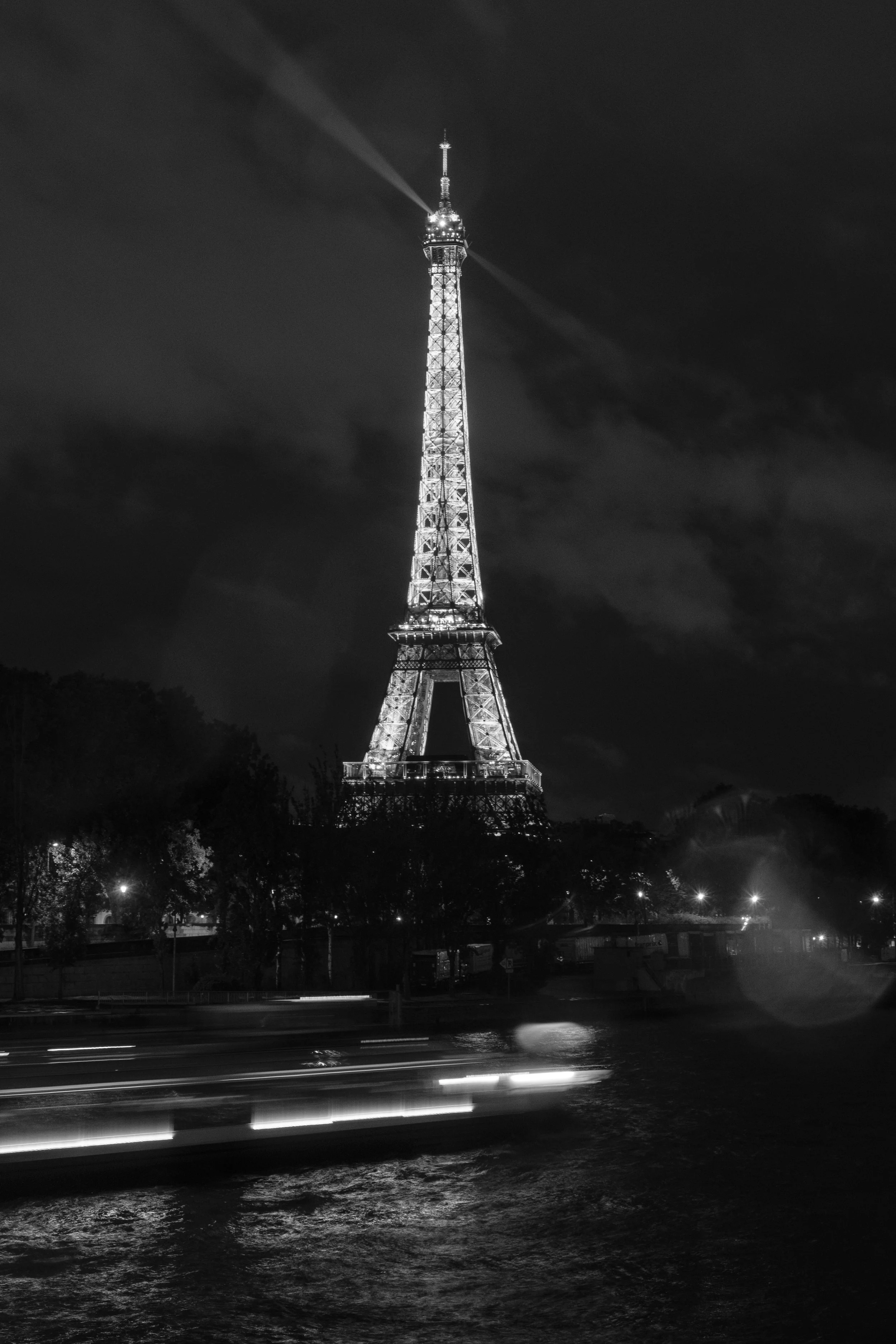 Der Eiffelturm bei Nacht, beleuchtet und mit Suchscheinwerfern, im Hintergrund dunkler Himmel mit Wolken, im Vordergrund Wasser mit Lichtspuren von Boote, in Schwarz-Weiß