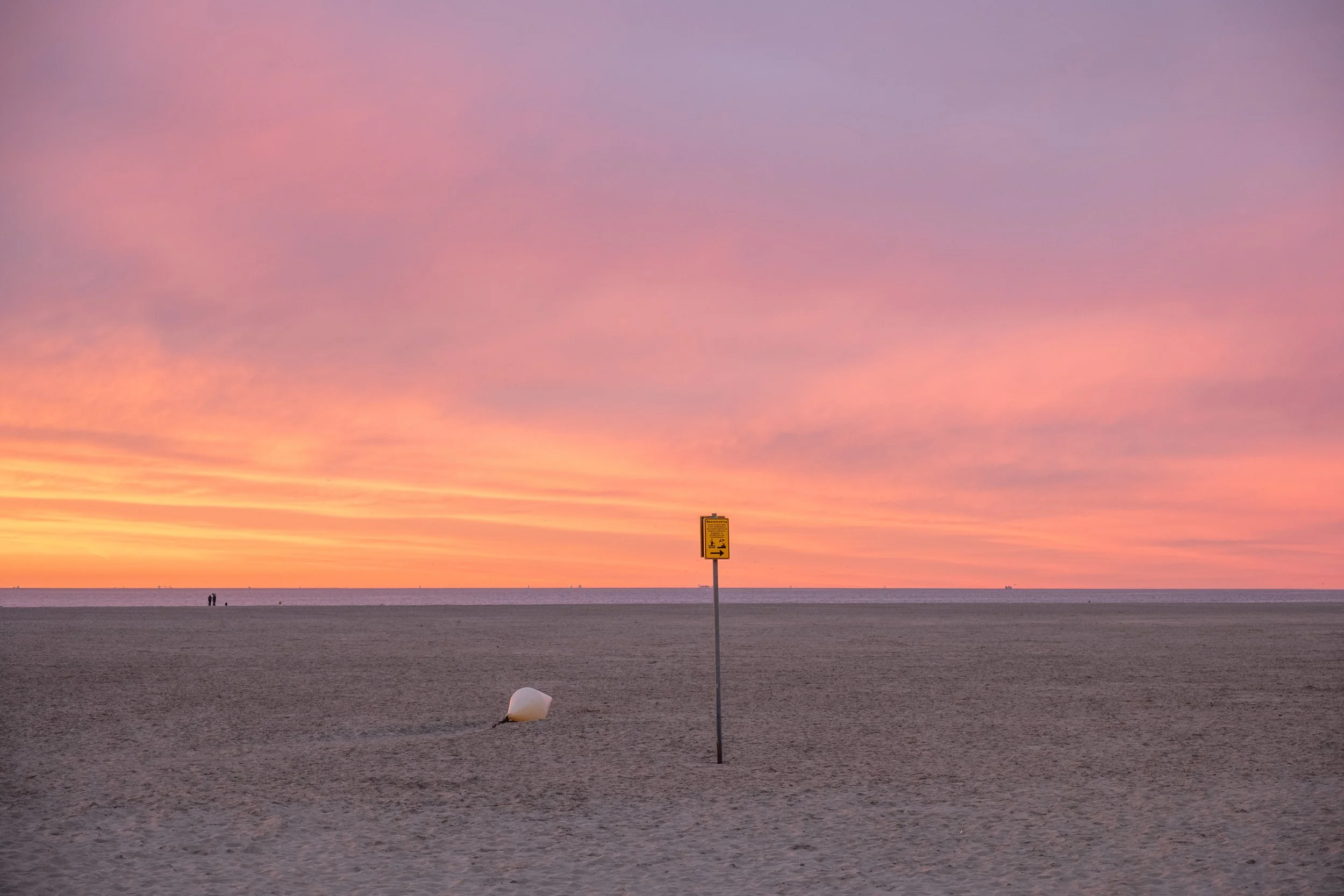 Ein Strand bei Sonnenuntergang mit einem gelben Warnschild, einem umgestürzten weißen Bötchen und zwei Menschen im Abstand zur linken Seite.