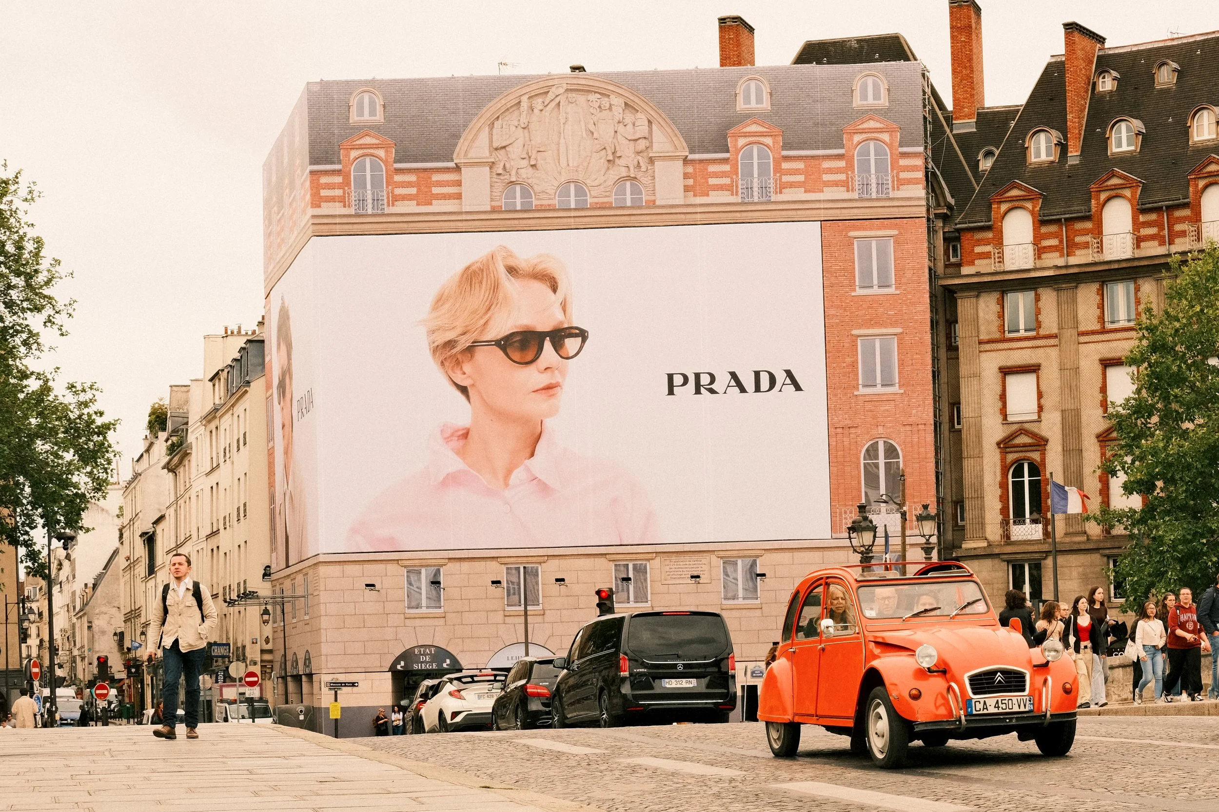 Straßenansicht in Paris mit einem roten Citroën 2CV, einem großen Prada-Werbebanner mit einer modischen Frau und mehreren Passanten am Tag.