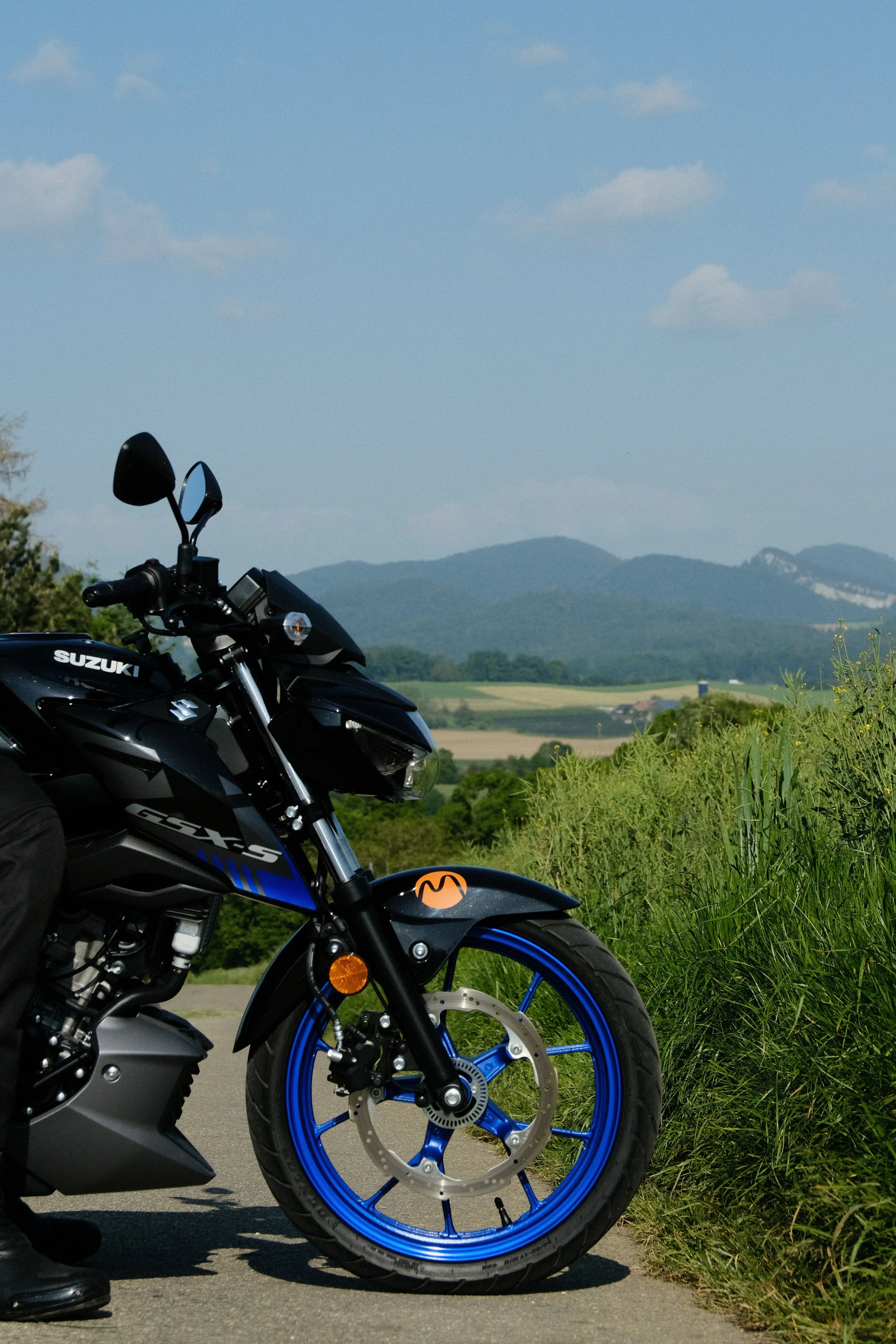 Schwarze Suzuki GSX-S Motorrad mit blauer Felge auf Landstraße mit grünen Wiesen und Hügel im Hintergrund.