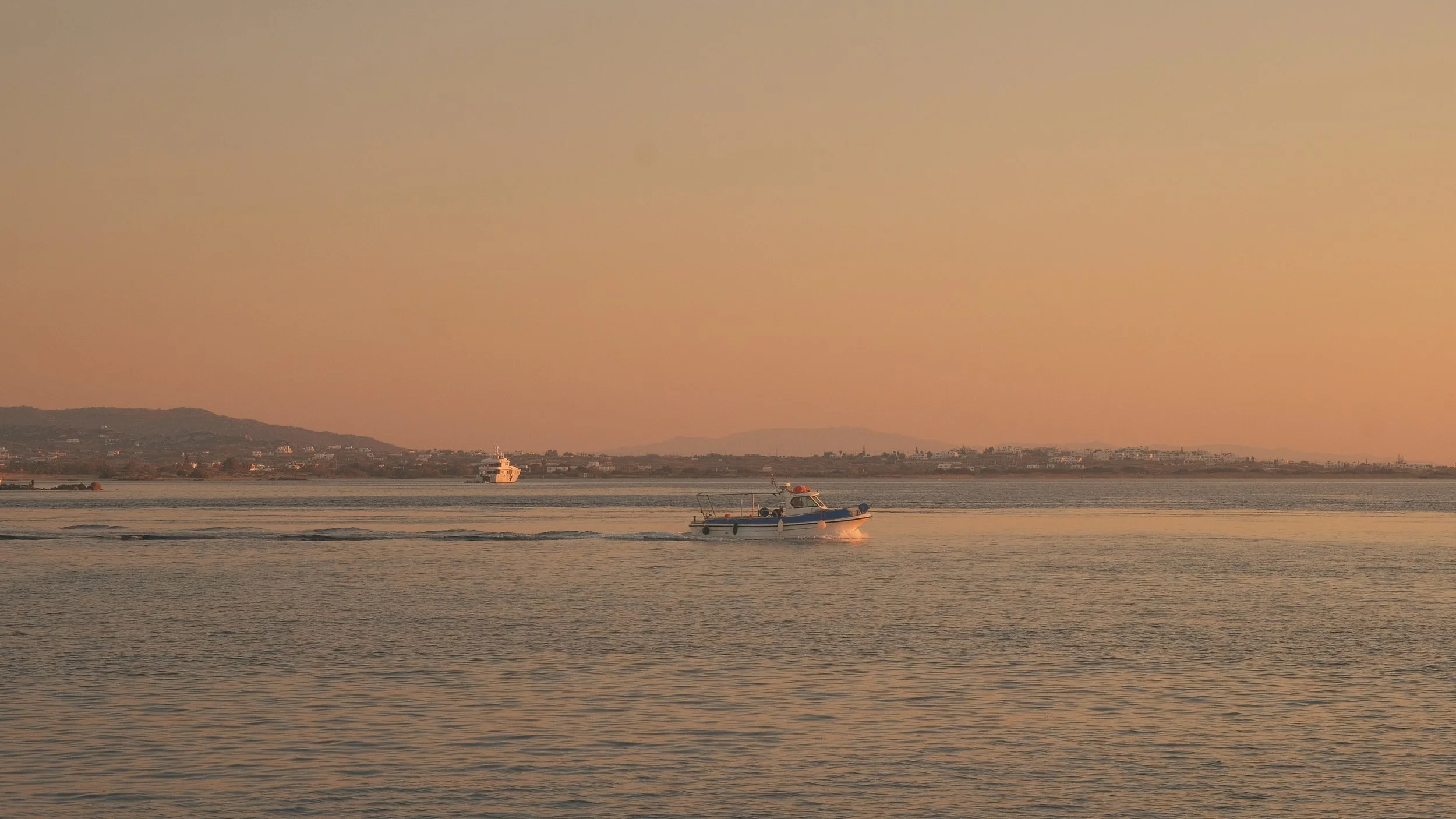 Ein Motorboot auf ruhigem Wasser während des Sonnenuntergangs, mit einer Küstenstadt im Hintergrund.