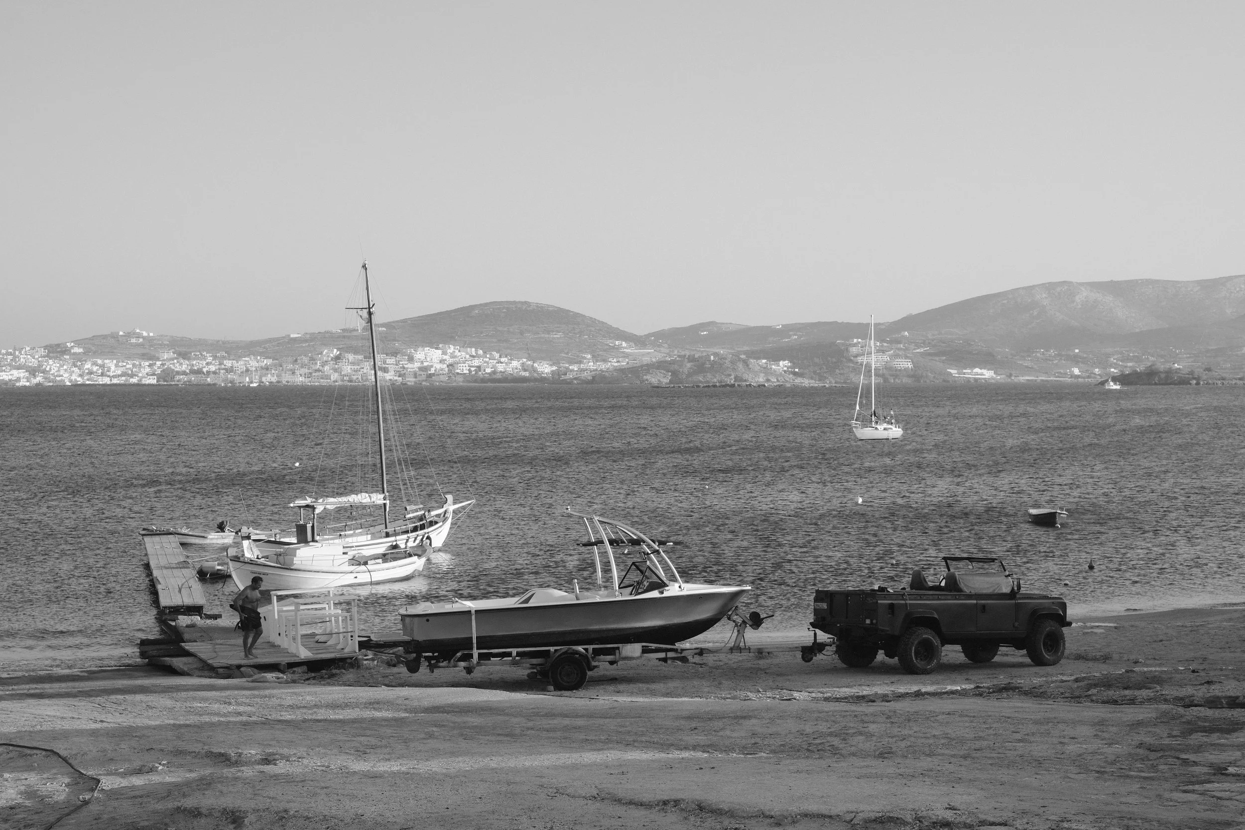 Schwarz-Weiß-Foto eines Hafens mit Booten, einem Lastwagen und einem Mann bei Sonnenuntergang am Wasser, mit einer Stadt und Hügel im Hintergrund.