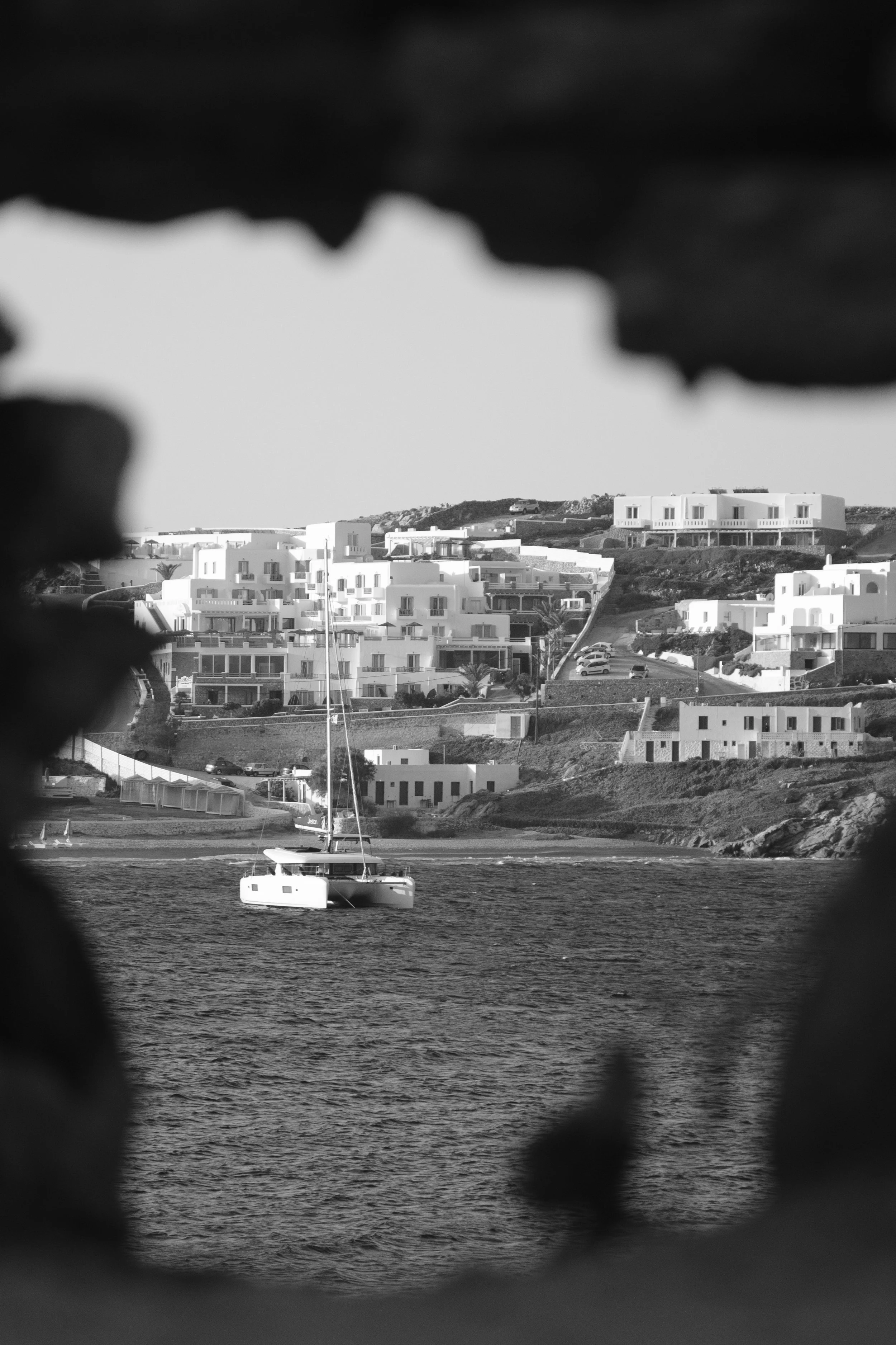 Ein Segelboot auf dem Wasser vor weißem Häuser in einer Küstenstadt, umrahmt von dunklen Blättern im Vordergrund, schwarz-weiß fotografiert.