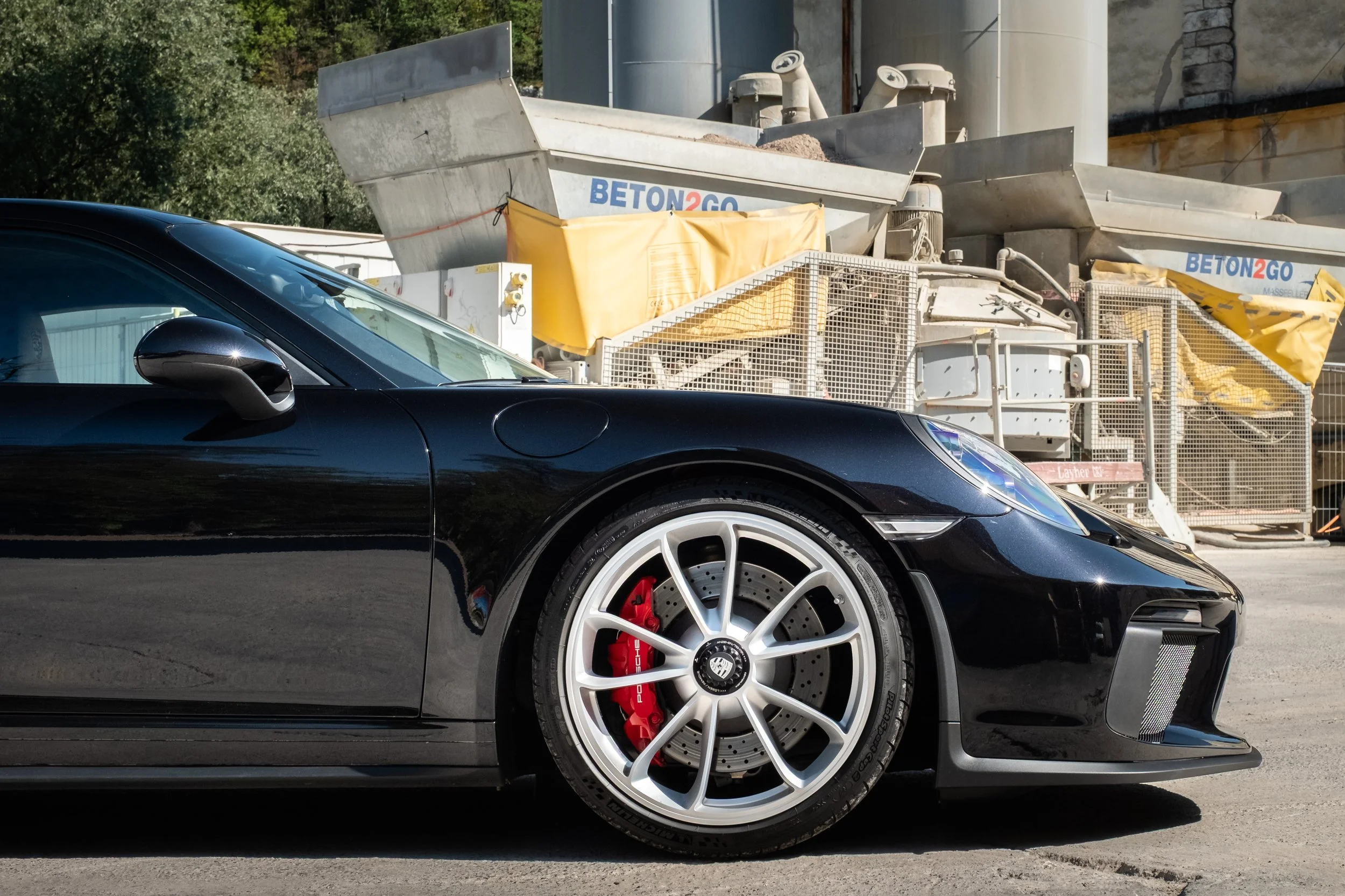 Schwarzer Sportwagen mit roten Bremsscheiben und silbernen Felgen, neben einem Baugerät mit Betonausleger, auf einer Baustelle.