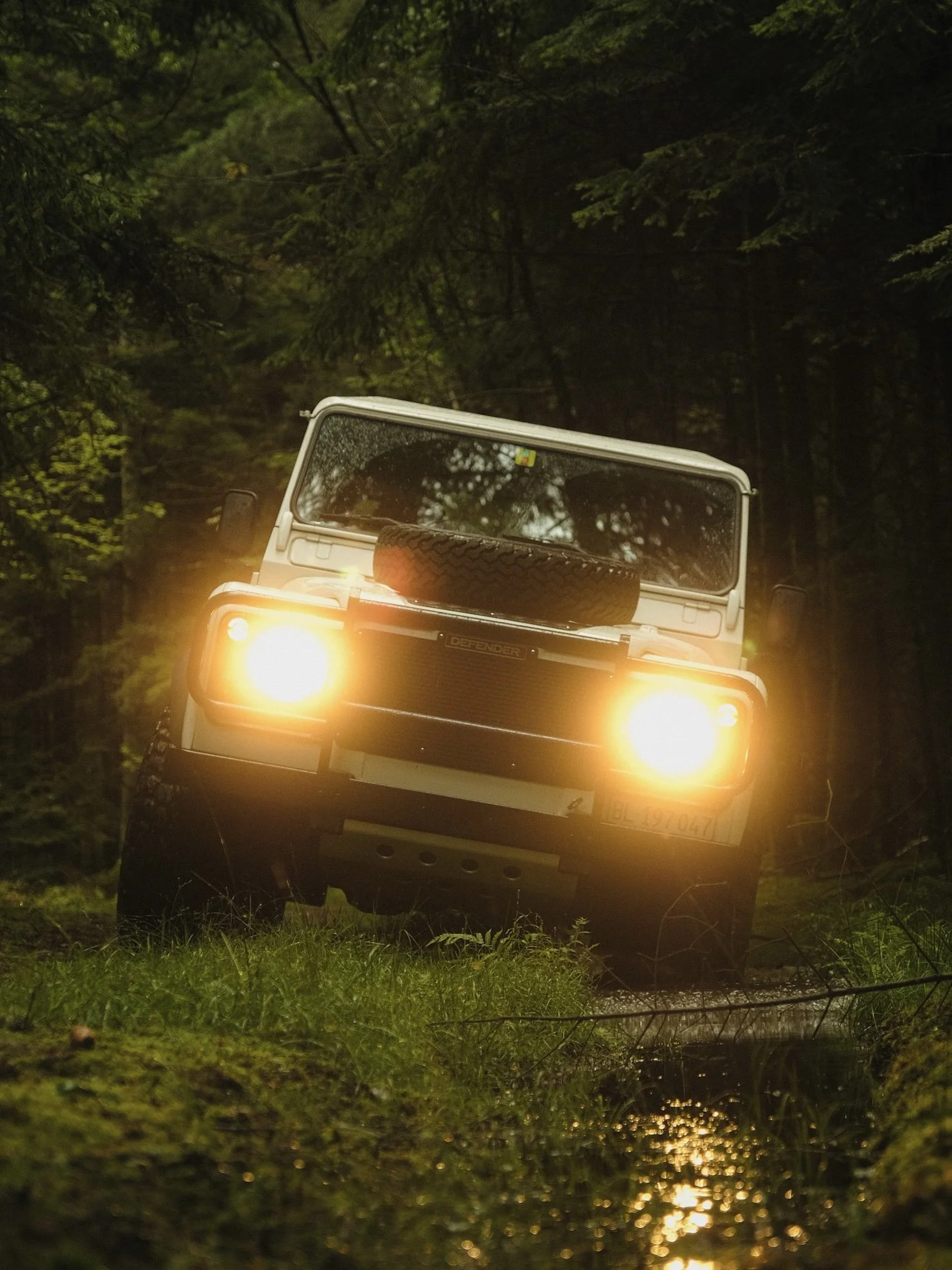 Ein weißes Geländefahrzeug mit eingeschalteten Scheinwerfern, das in einem Waldstück auf einem matschigen Pfad unterwegs ist.