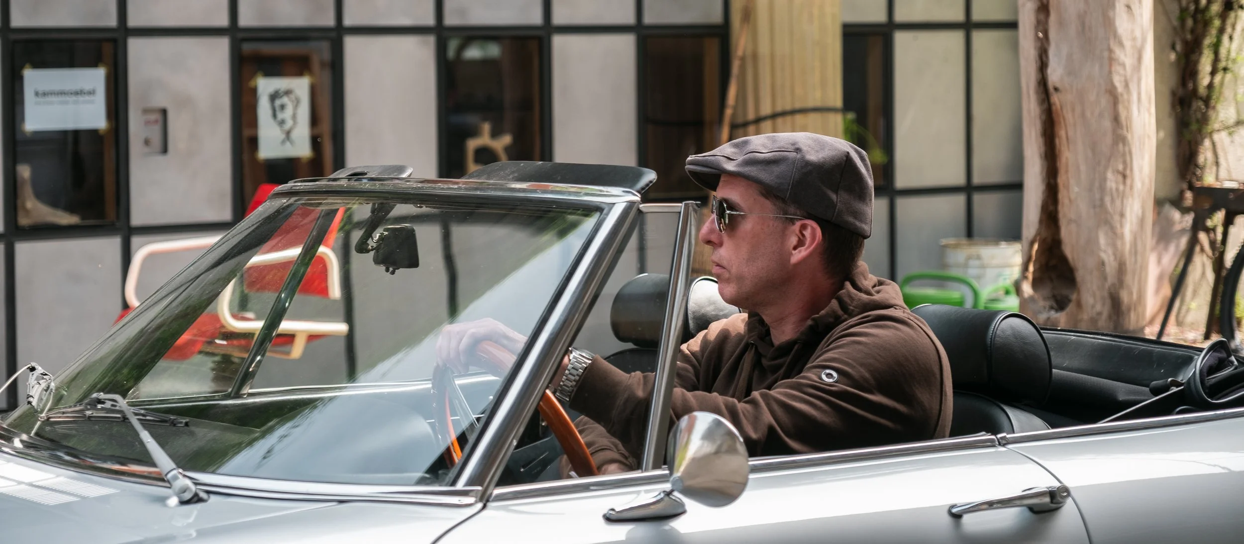 Ein Mann sitzt in einem silbernen Cabriolet, trägt eine braune Kapuzenjacke, eine dunkle Kappe und Sonnenbrille, während er am Steuer sitzt.