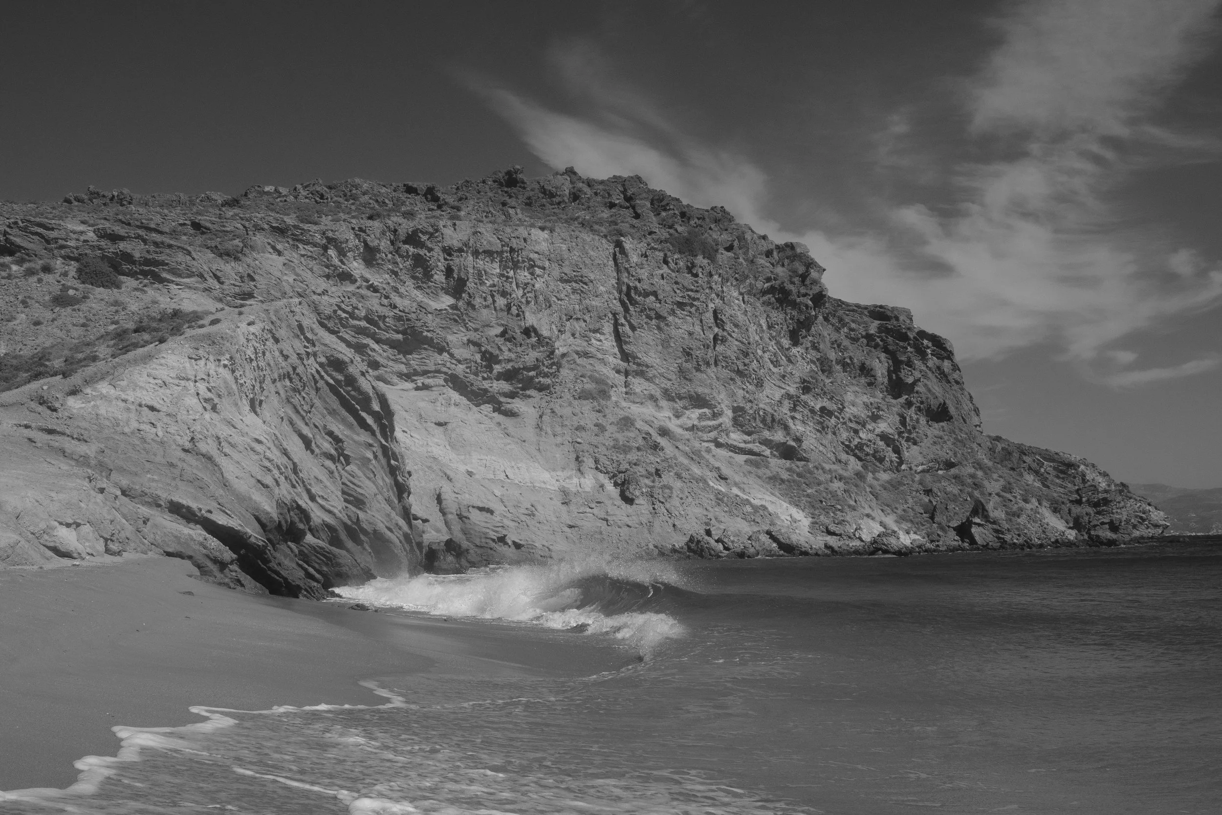 Schwarz-weiß Foto eines Strandes mit Klippen im Hintergrund und Wellen, die an den Strand schlagen.