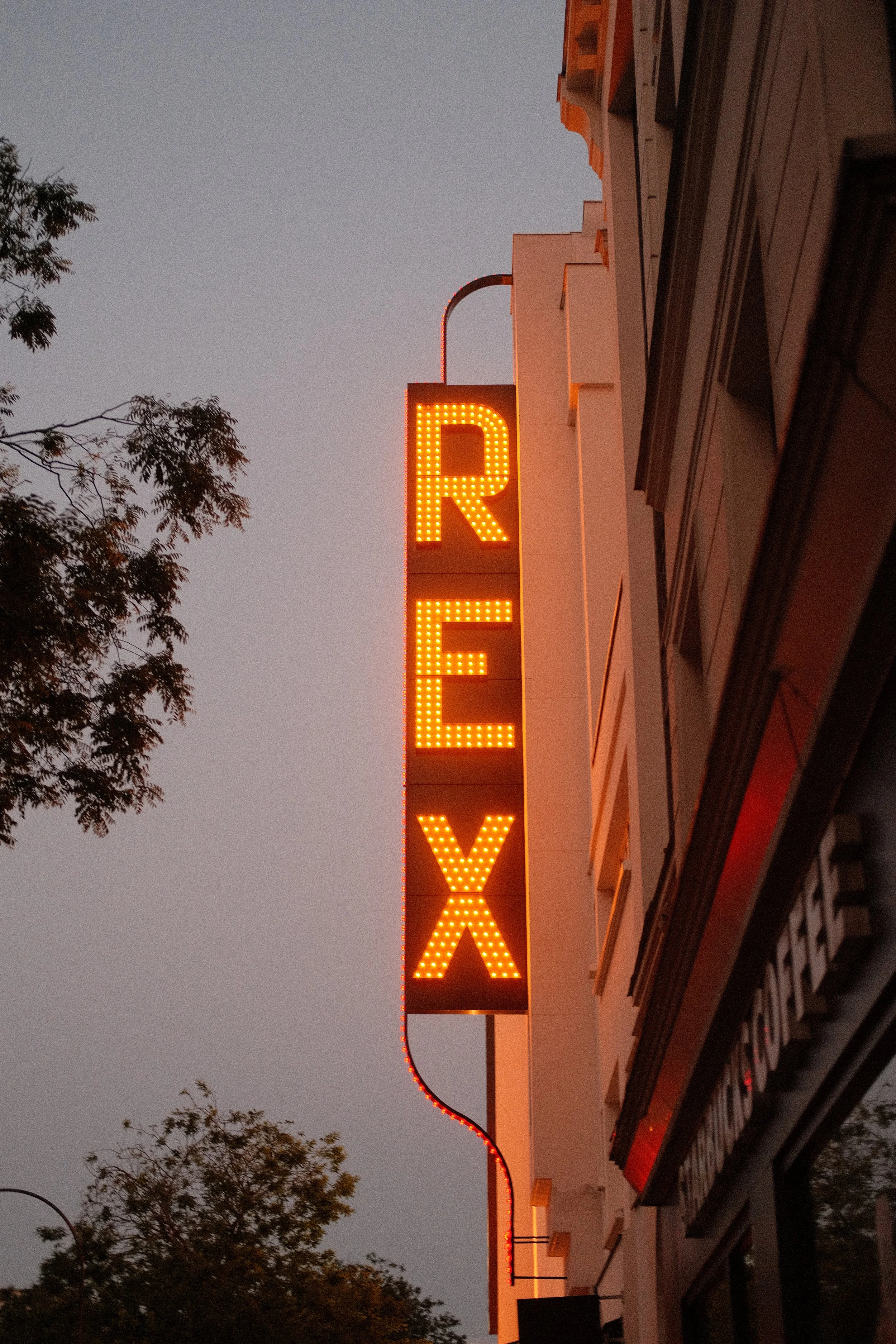 Das Bild zeigt ein leuchtendes Neon-Schild mit den Buchstaben R, E, V, X in orangefarbenen Lichtern an der Seite eines Gebäudes, umgeben von Bäumen bei Dämmerung.