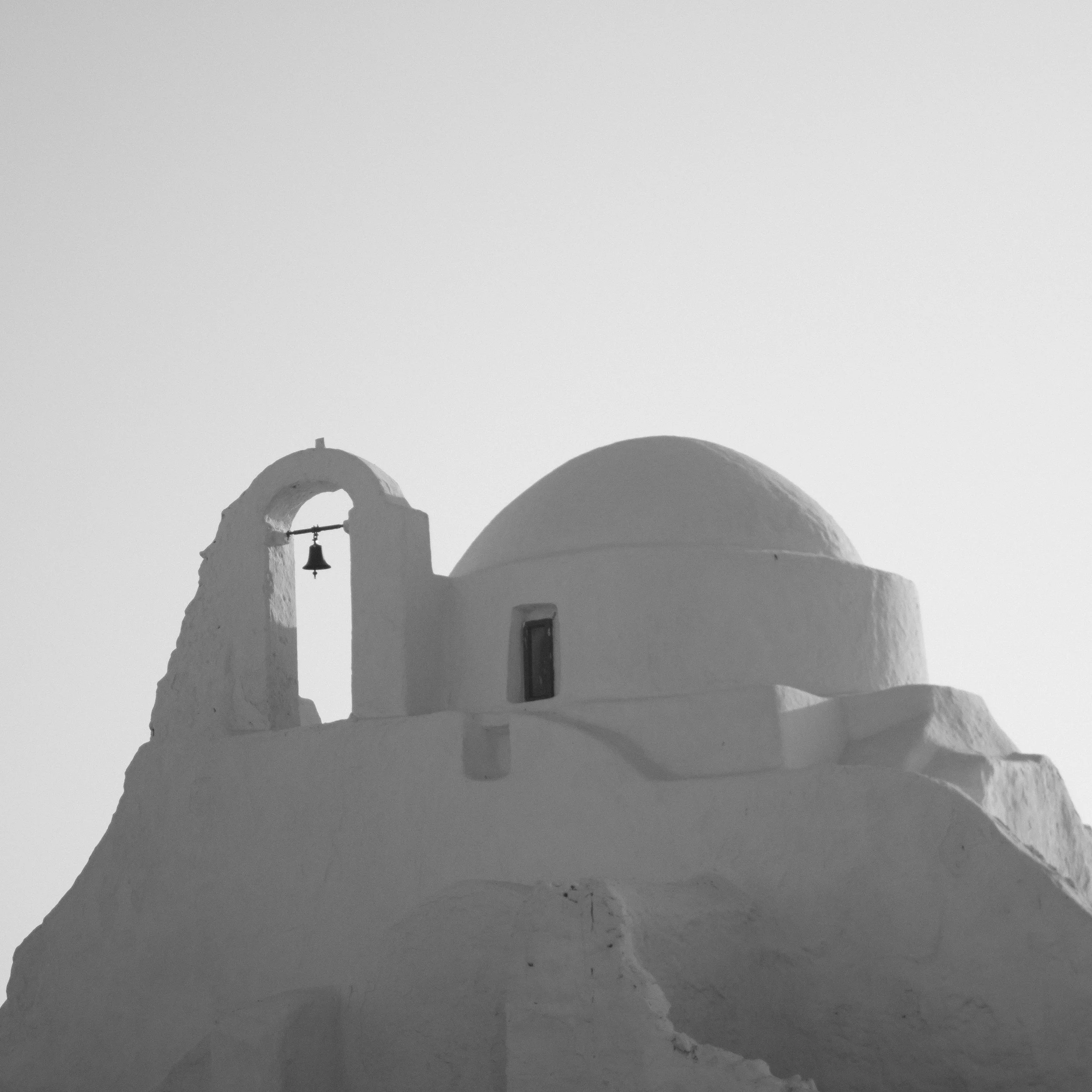 Weiße Kirche mit Kuppel und Glocke auf einem Hügel, typisch für griechische Architektur, vor hellem Himmel.