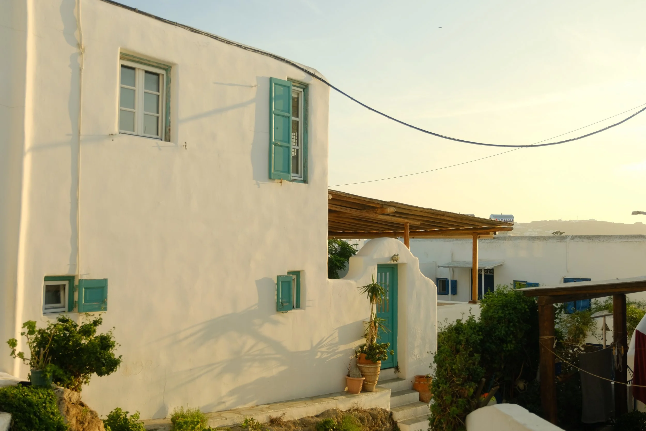 Weiße Hausfassade mit blauen Fensterläden und Tür, Potted Plants und einem Holzdach im Sonnenlicht