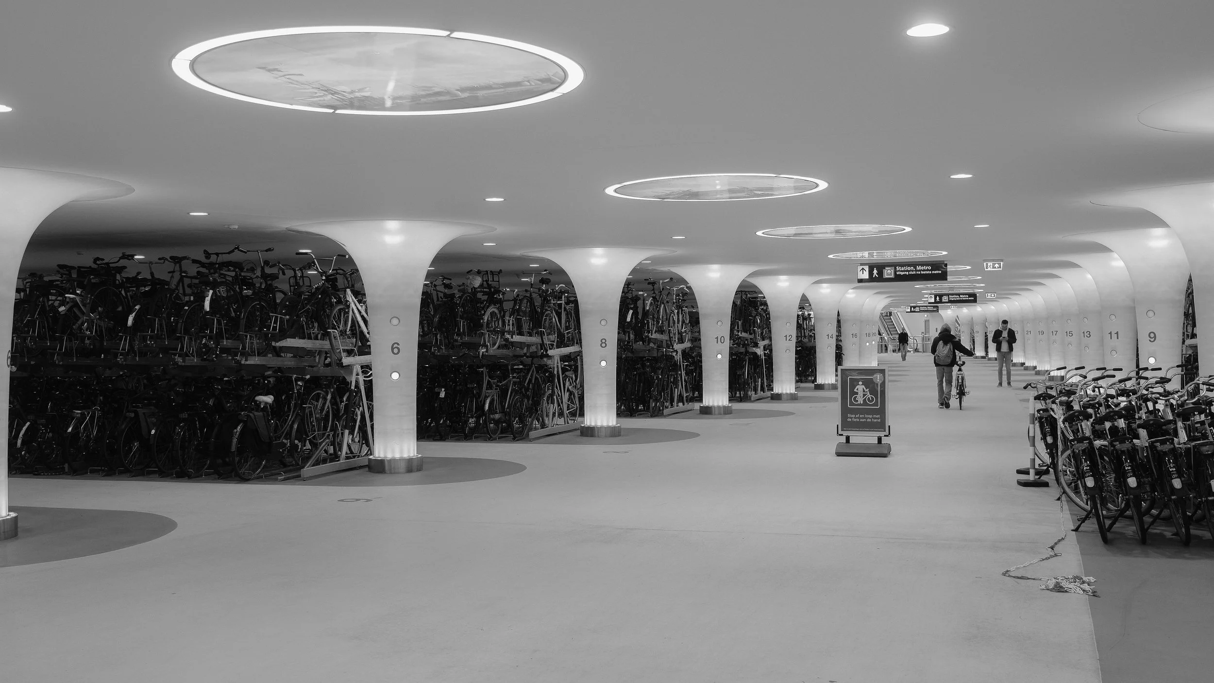 Innenaufnahme eines modernen Fahrradparkhauses mit mehreren Fahrradständern und Fahrrädern, einigen Personen, die Fahrrad fahren oder gehen, sowie Hinweisschilder an der Decke.