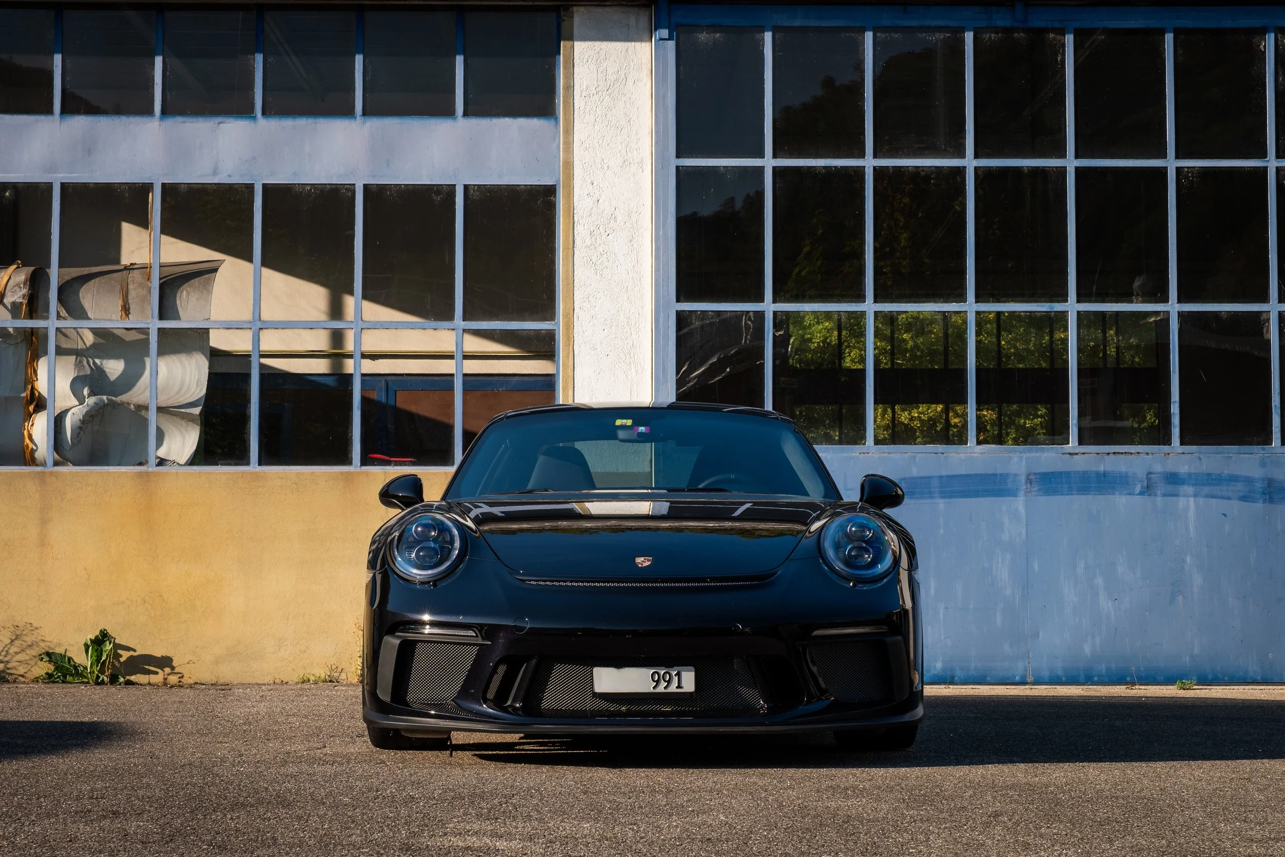 Schwarzer Porsche 911 vor einer alten Fabrik mit großen Fensterflächen.