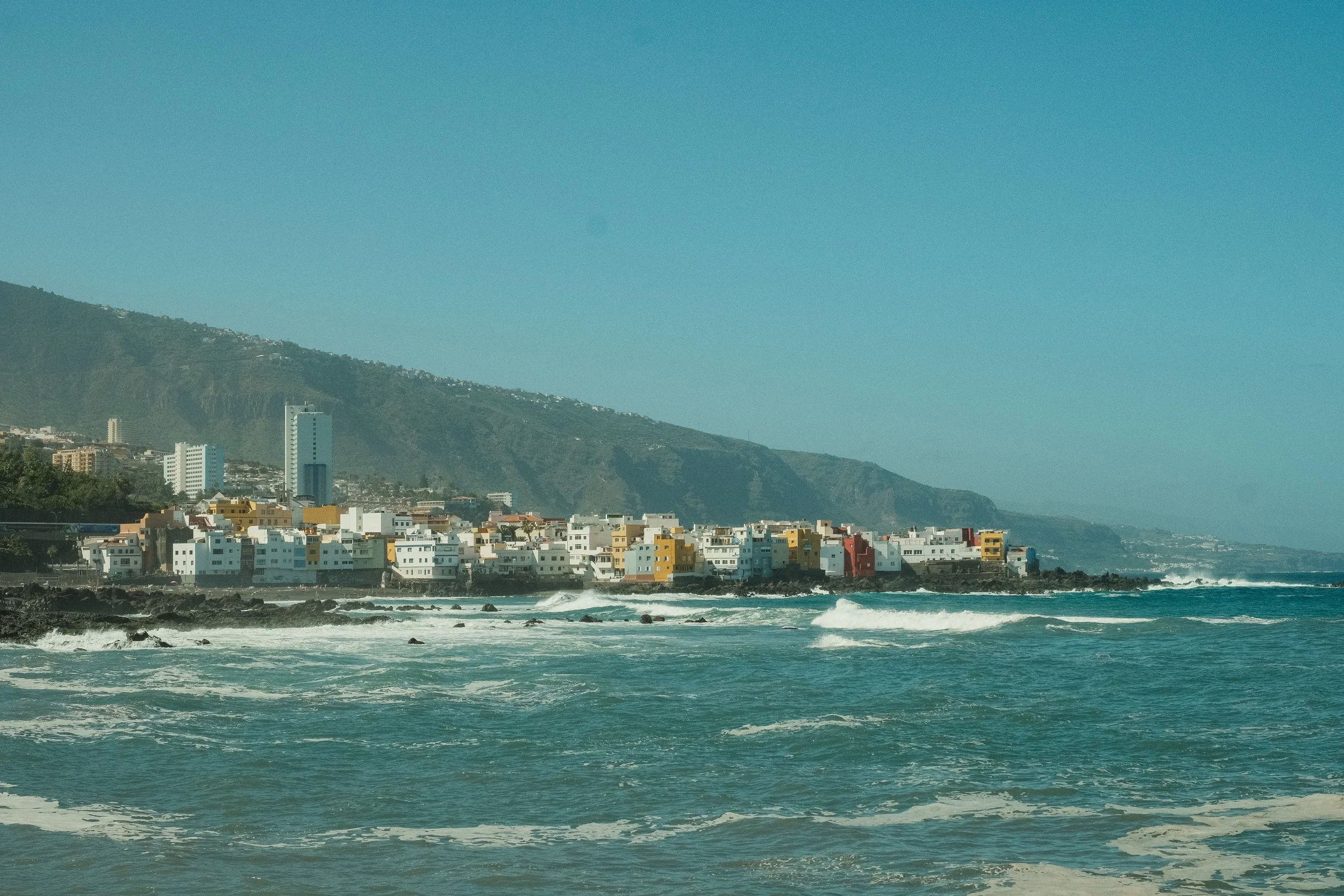 Blick auf eine Küstenstadt mit bunten Häusern am Meer, Berge im Hintergrund, klarem Himmel und Wellen im Vordergrund.