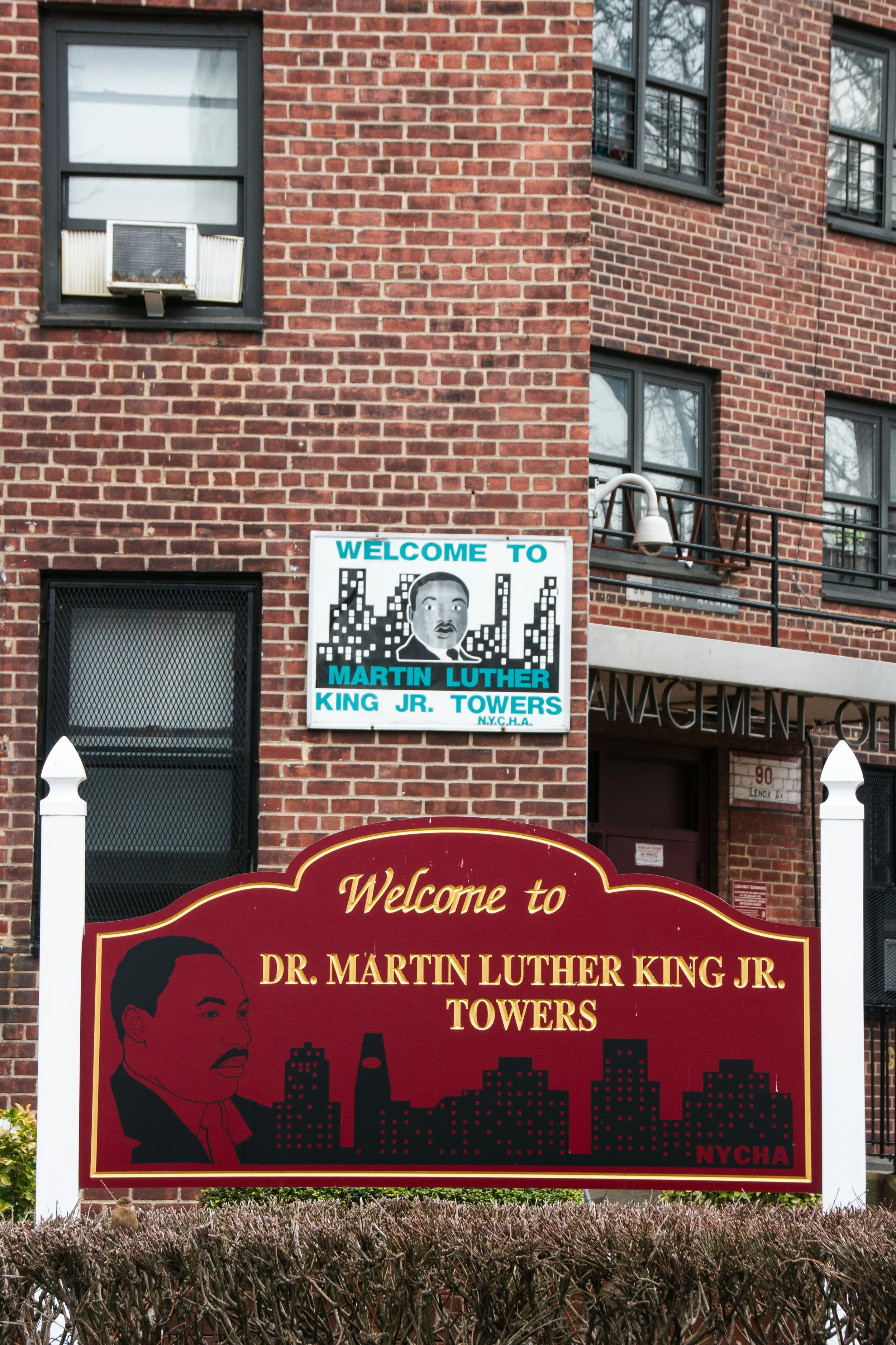 Ein rotes Willkommensschild vor einem roten Backsteingebäude mit mehreren Fenstern und einem aufgestellten Gemälde. Das Schild begrüßt Besucher bei den 'Dr. Martin Luther King Jr. Towers' in New York City, mit einer Porträtillustration von Martin Lut