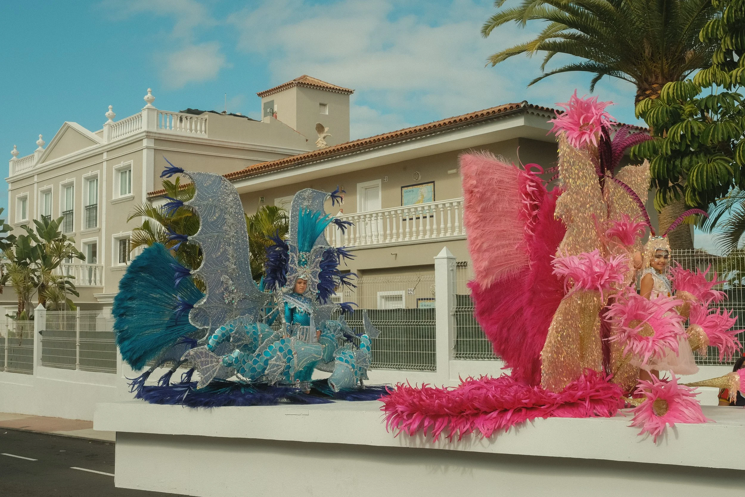 Blaue und pinke Karnevalskostüme auf einem Faschingswagen, im Hintergrund weiße Gebäude mit Palmen, bei schönem Wetter.