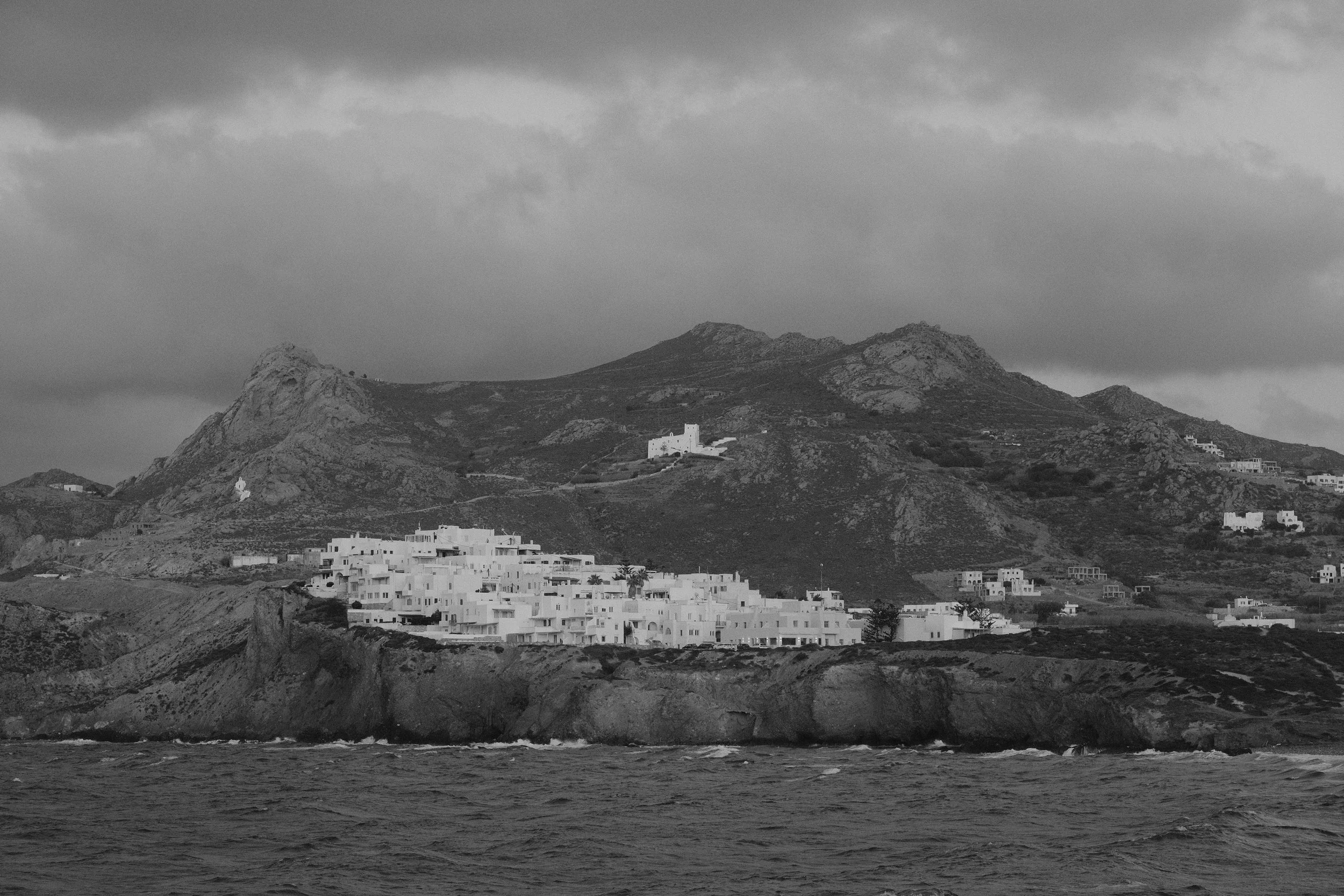 Schwarze und weiße Küstenstadt mit weißen Gebäuden auf einer Klippe, umgeben von Bergen und dunklen Wolken am Himmel.