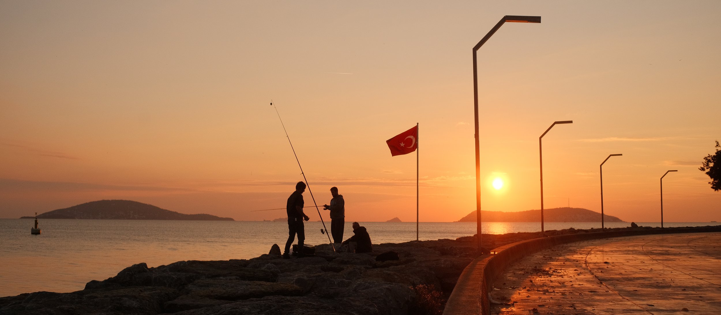 Sonnenuntergang am Meer mit Silhouetten von drei Menschen, die Angeln im Vordergrund, türkische Flagge, Leuchtturm, kurvige Strandpromenade und entfernte Inseln im Hintergrund.