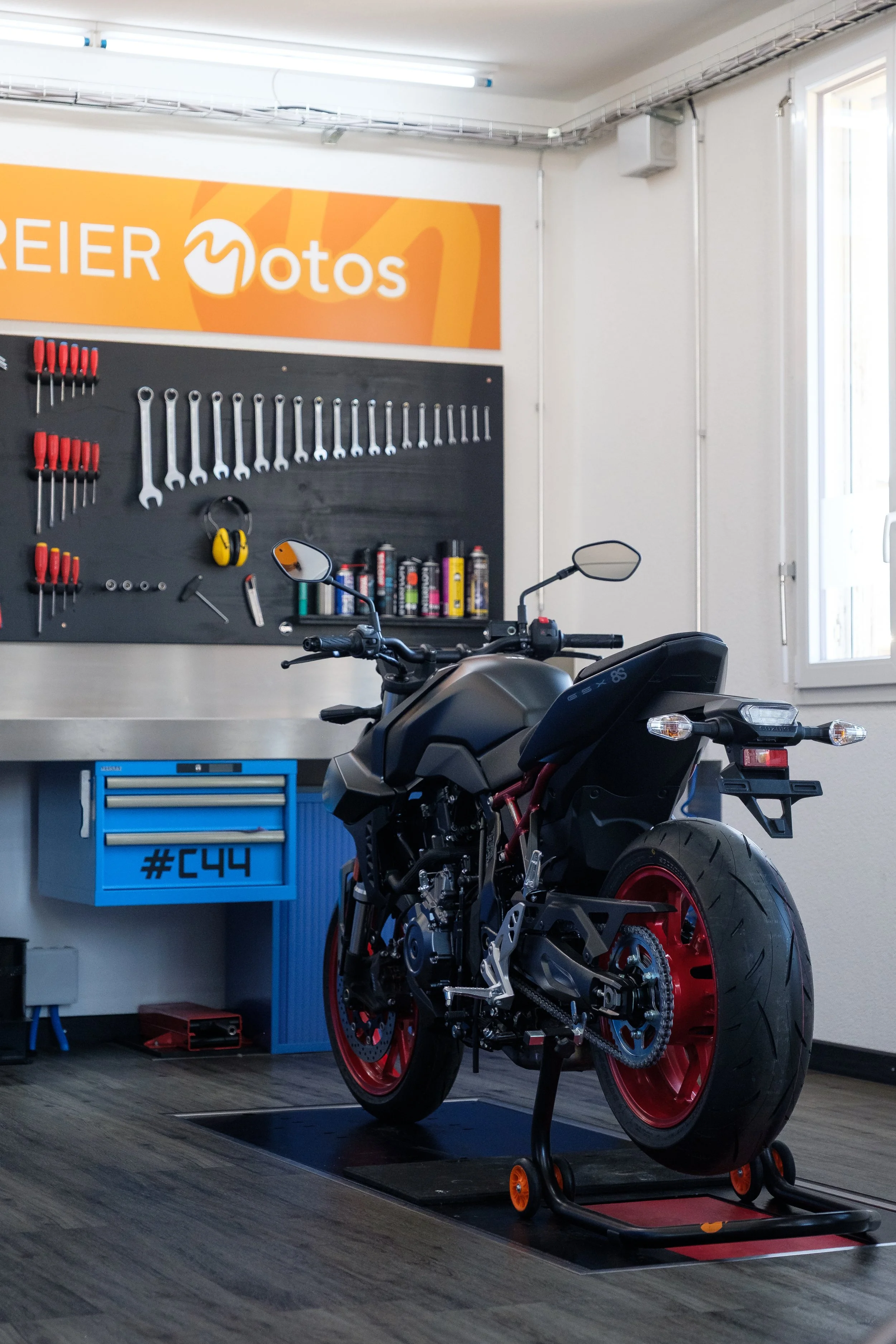 Ein Motorrad in einer Werkstatt auf einem Montageständer, im Hintergrund eine Werkzeugtafel mit Schraubenziehern und Schraubenschlüsseln sowie eine Arbeitsplatte mit Werkzeugen und Pflegeprodukten.