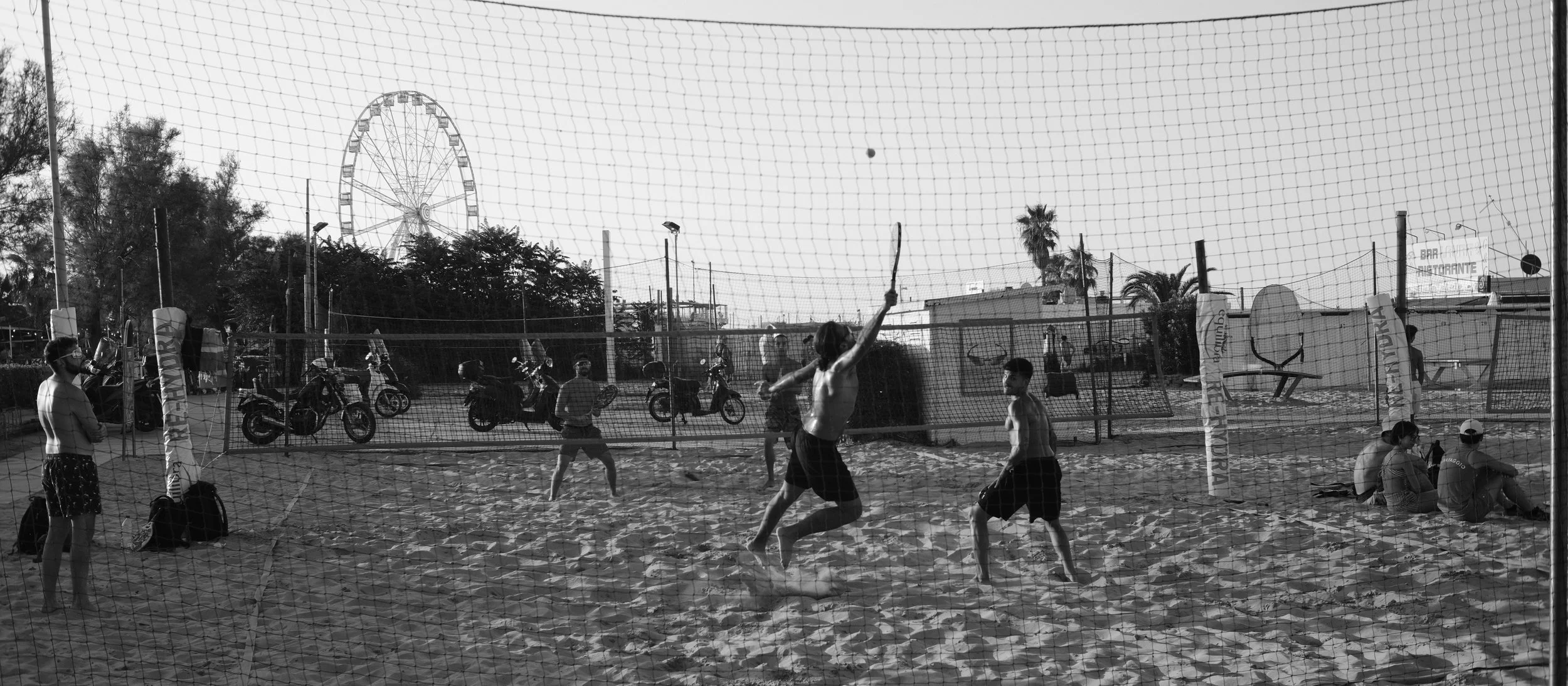 Jugendliche spielen Beachvolleyball auf einem Sandplatz, während andere Jugendliche und Erwachsene im Hintergrund amüsiert sind. Es gibt einen Riesenrad im Hintergrund und Motorräder entlang der Zaunlinie.