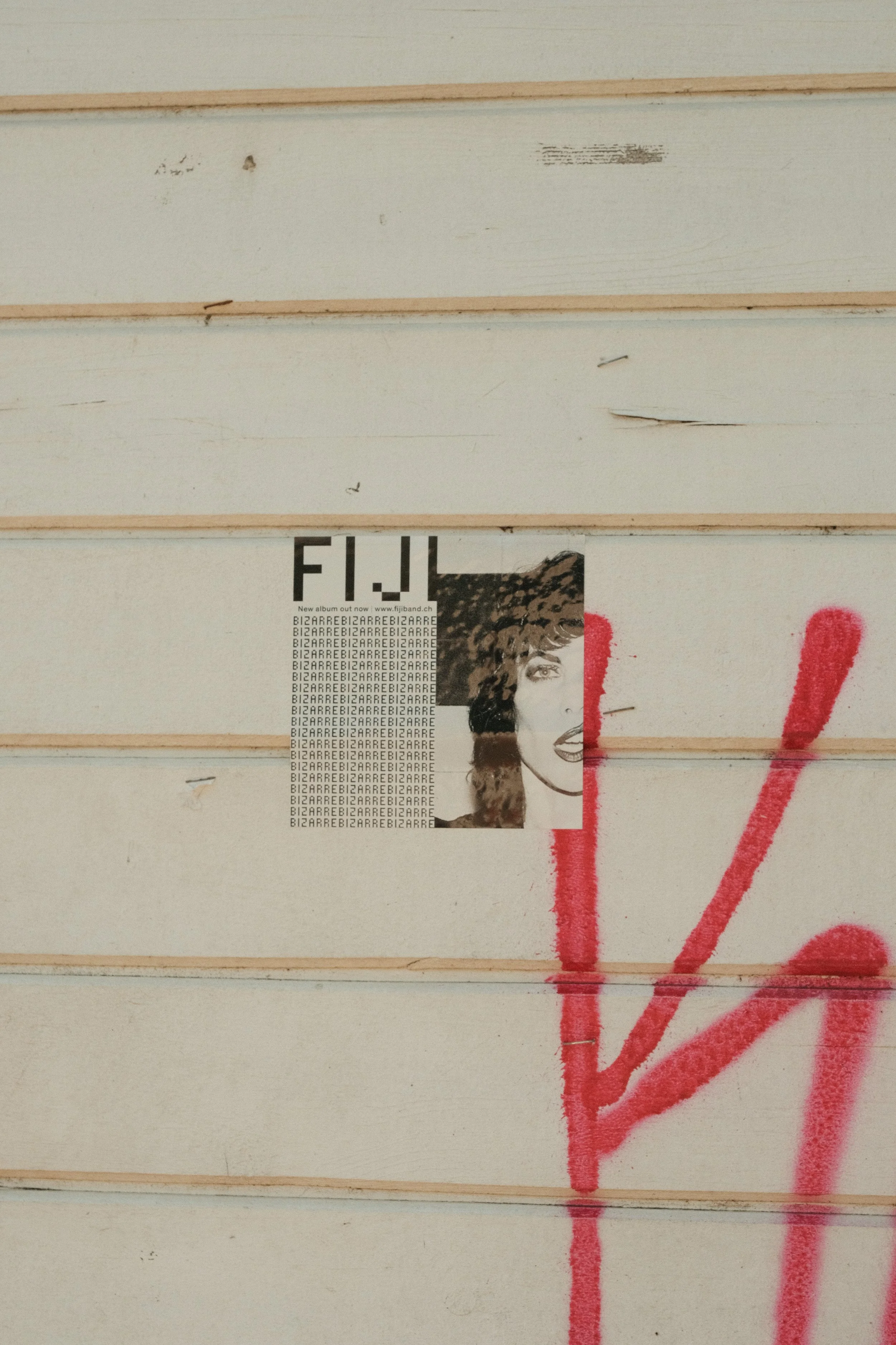 Wand mit Graffiti und einem Plakat einer Frauenzeichnung, die teilweise verdeckt ist, sowie roter Sprayfarbe. Die Wand hat horizontal verlaufende Holzpaneele.