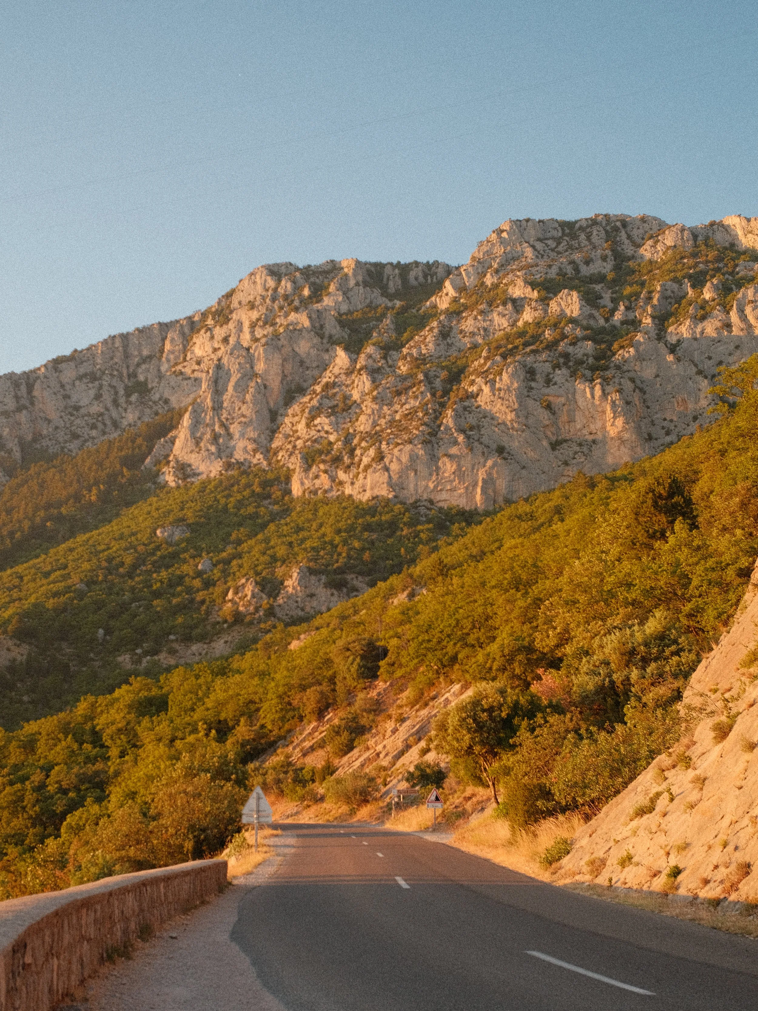 Eine kurvige Straße, die durch eine bergige Landschaft führt, mit Bäumen und Felsen bei Sonnenuntergang.