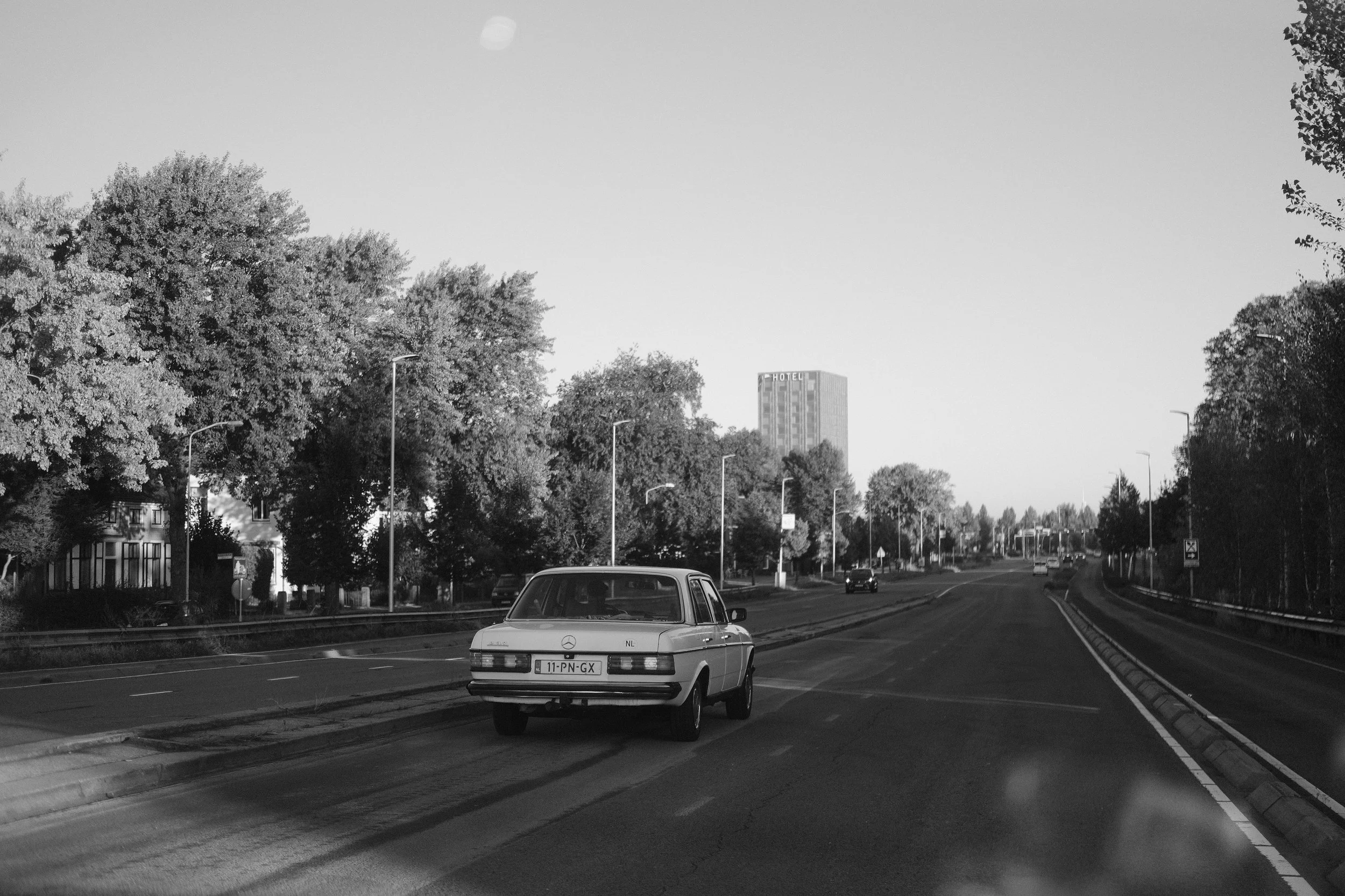 Ein schwarzes und weißes Foto einer Straße mit einem weißen alten Mercedes-Benz Auto, Bäumen auf der linken und rechten Seite, im Hintergrund ein Hotel und andere Fahrzeuge auf der Straße.
