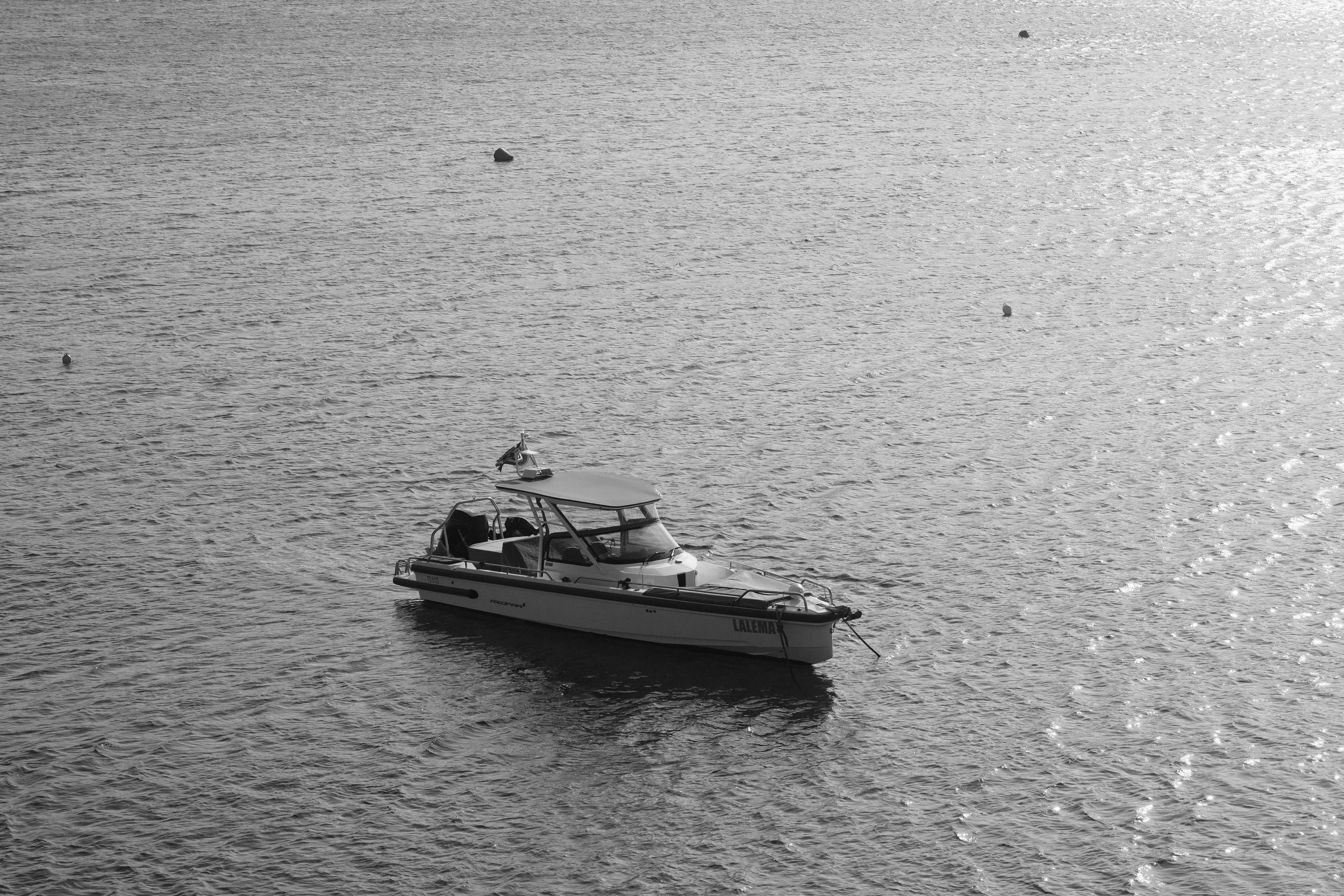 Ein Boot schwimmt auf einer ruhigen Wasseroberfläche, umgeben von kleinen Bojen. Das Bild ist in Schwarz-Weiß aufgenommen.