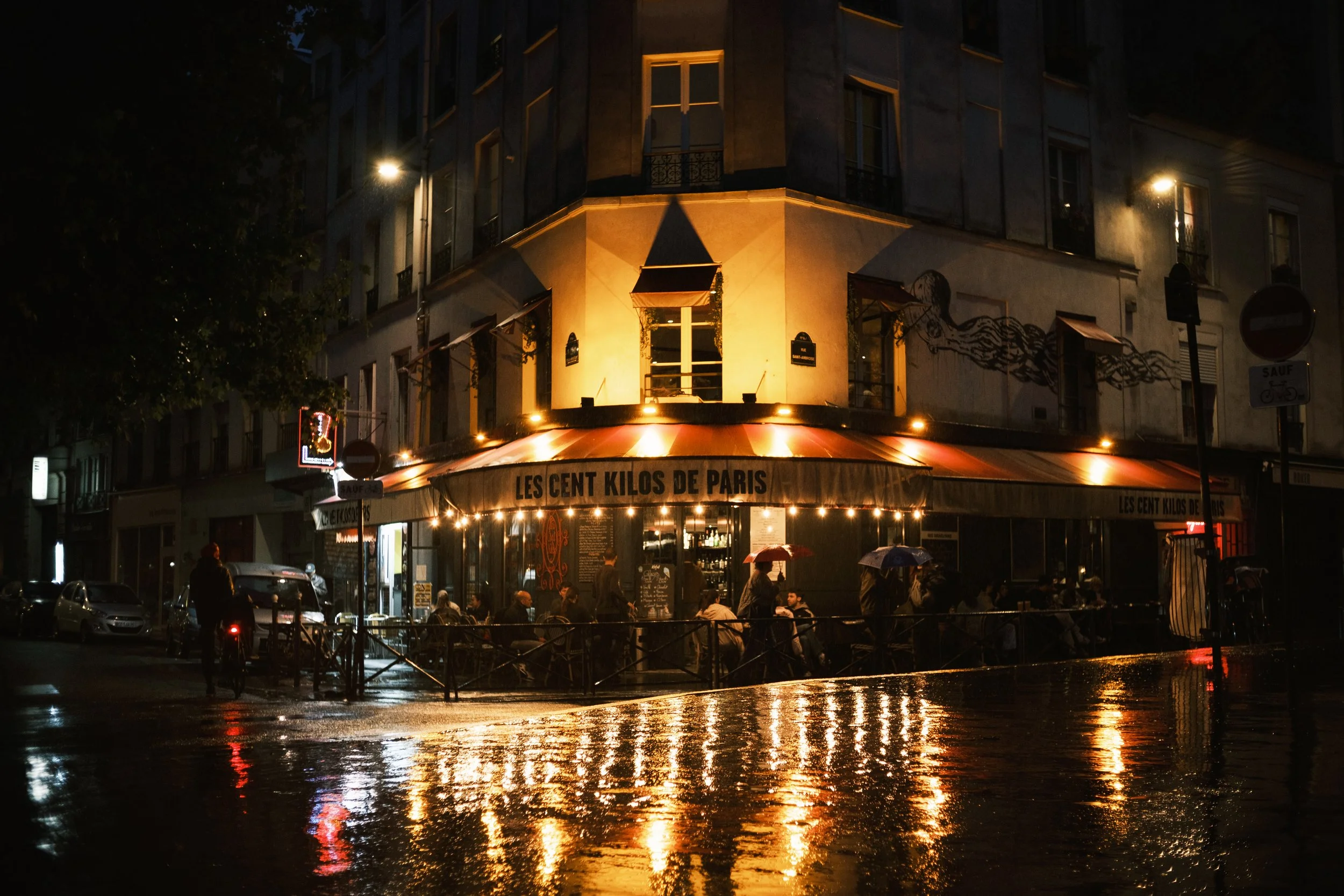 Das Bild zeigt ein Cafe in Paris namens 'Les Cent Kilos de Paris' bei Nacht. Das Cafe ist beleuchtet mit warmem Licht, Leute sitzen draußen unter Regenschirmen, Regen spiegelt sich auf der Straße, im Hintergrund sind Gebäude und Straßenlaternen sicht