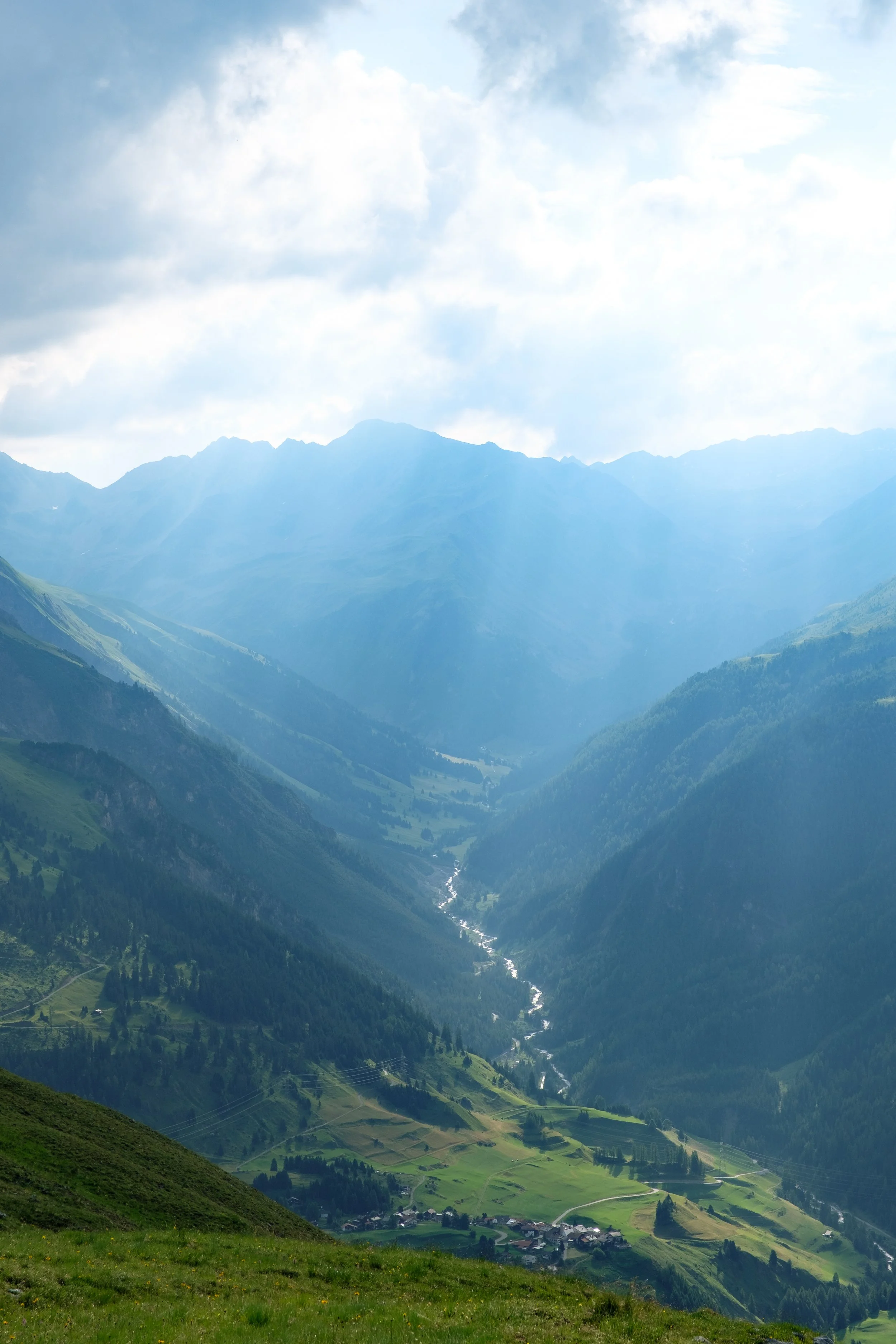 Berglandschaft mit einem Tal, einem Fluss und kleinen Häusern, umgeben von grünen Hügeln und hohen Bergen im Hintergrund, unter einem bewölkten Himmel.