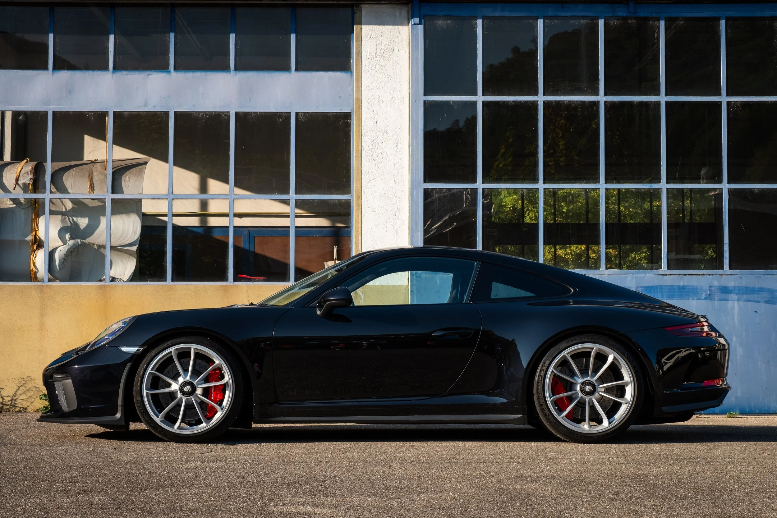 Schwarzer Sportwagen, vermutlich ein Porsche, parkend vor einem Industriegebäude mit großen Fenstern.