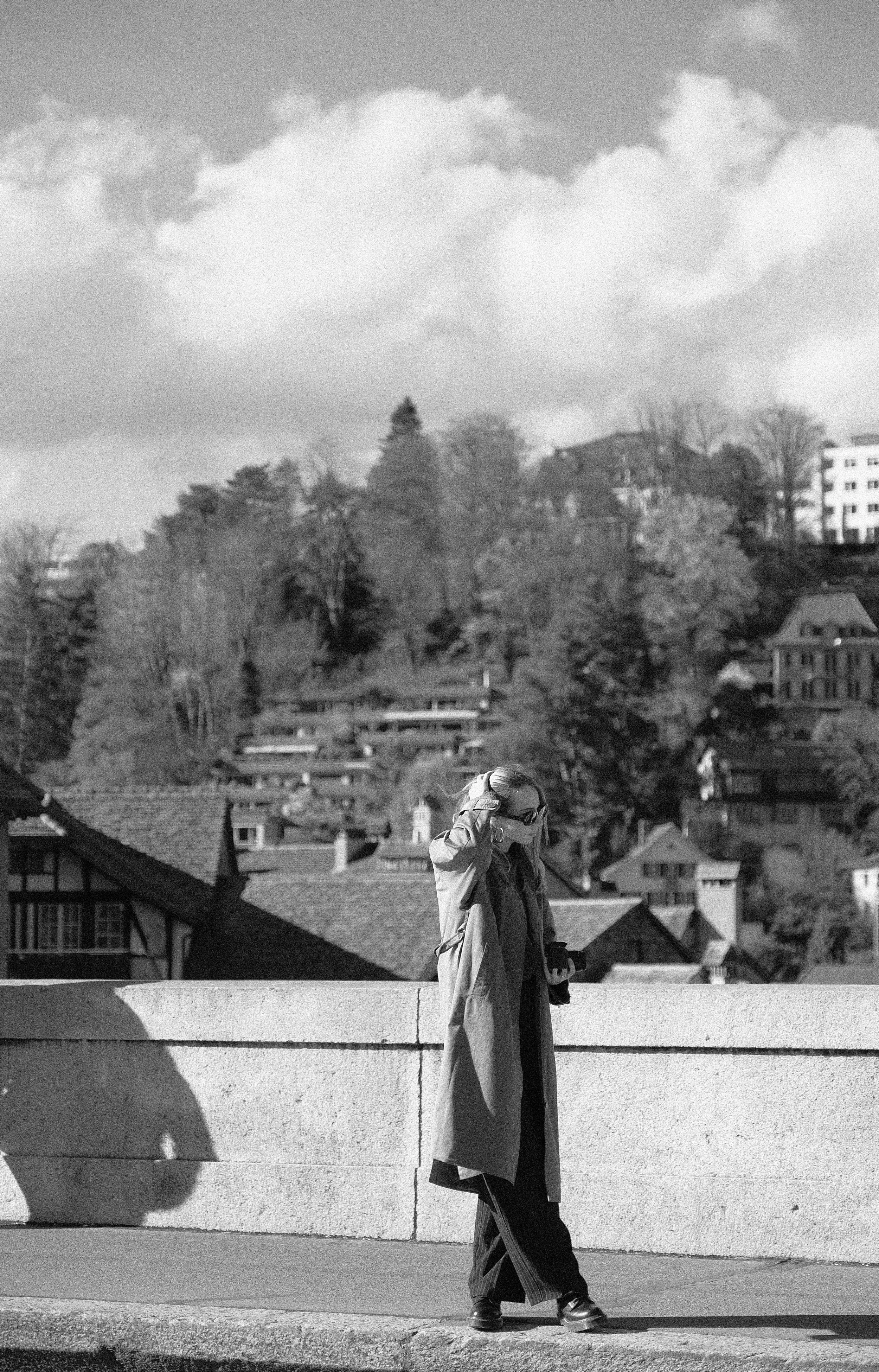 Frau geht auf einer Brücke mit Blick auf städtische Häuser und Bäume im Hintergrund, während sie eine Sonnenbrille trägt und ein Smartphone hält.