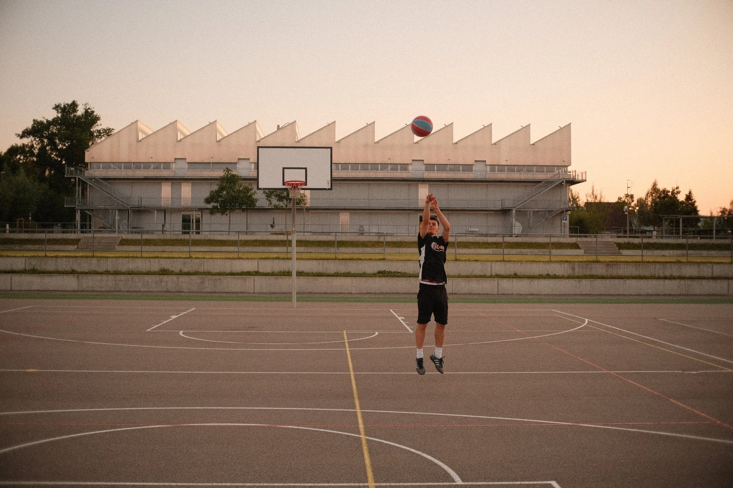 Ein Teenager wirft einen Basketball auf einem leeren Basketballplatz bei Sonnenuntergang. Im Hintergrund ist eine moderne Gebäudearchitektur zu sehen.