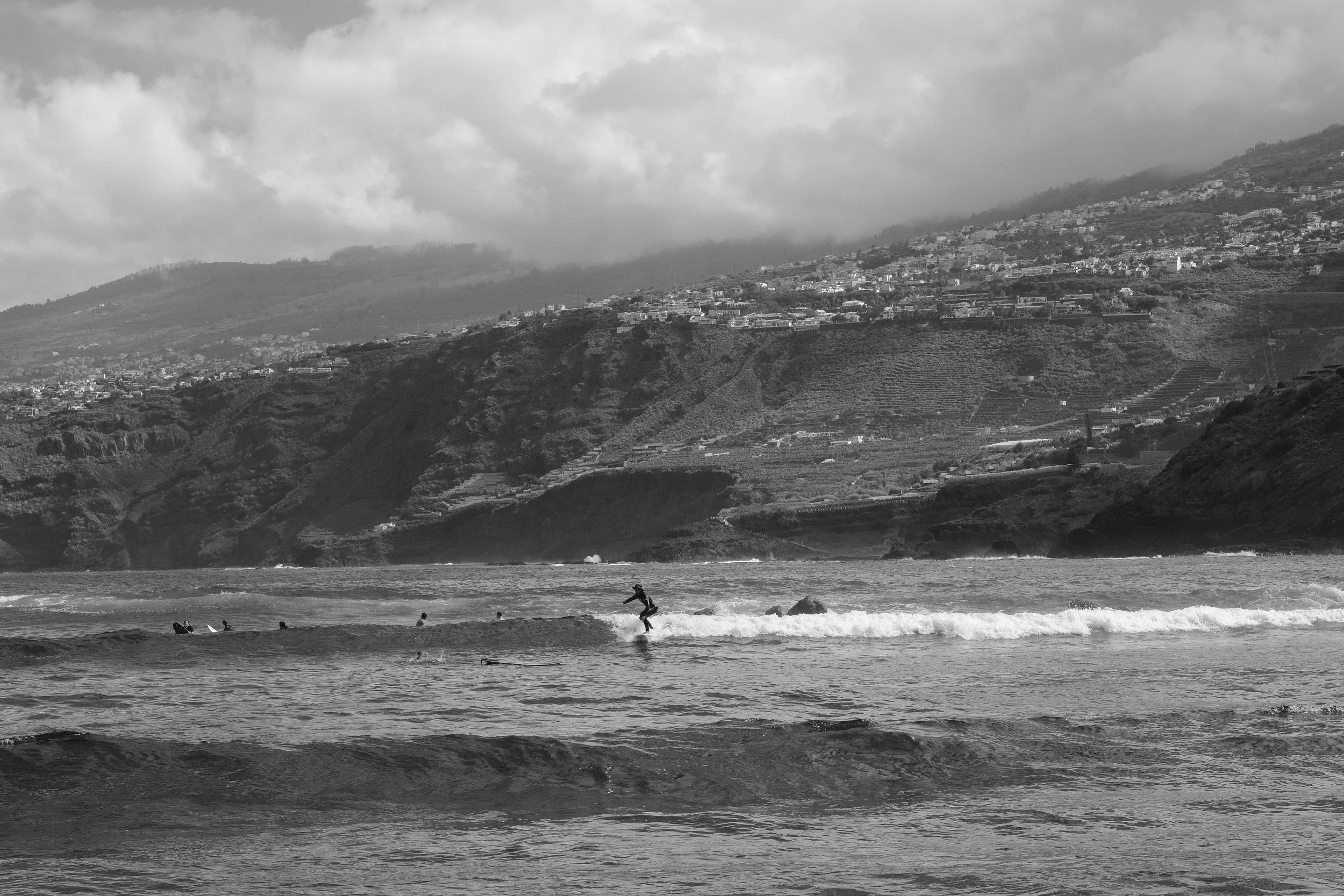 Strand mit Wellen, Surfer im Wasser und eine Person beim Surfen, im Hintergrund steile Hügel mit Häusern und bewässerten Terrassen, bewölkter Himmel.