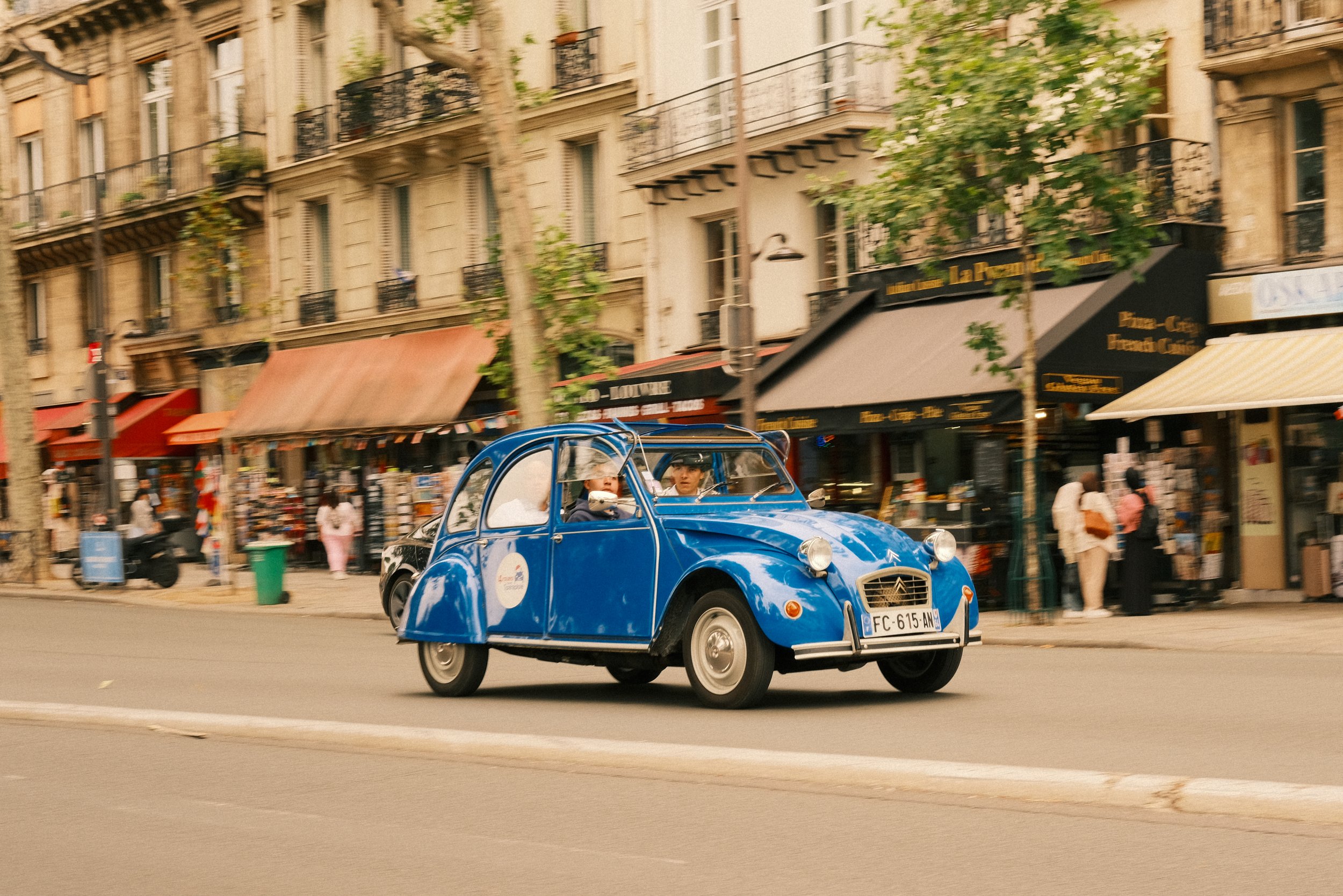 Ein blaues Citroën 2CV Auto fährt auf einer Stadtstraße vorbei an Geschäften und Passanten. Das Auto trägt Fahrer und Beifahrer, die beide in die Kamera schauen.