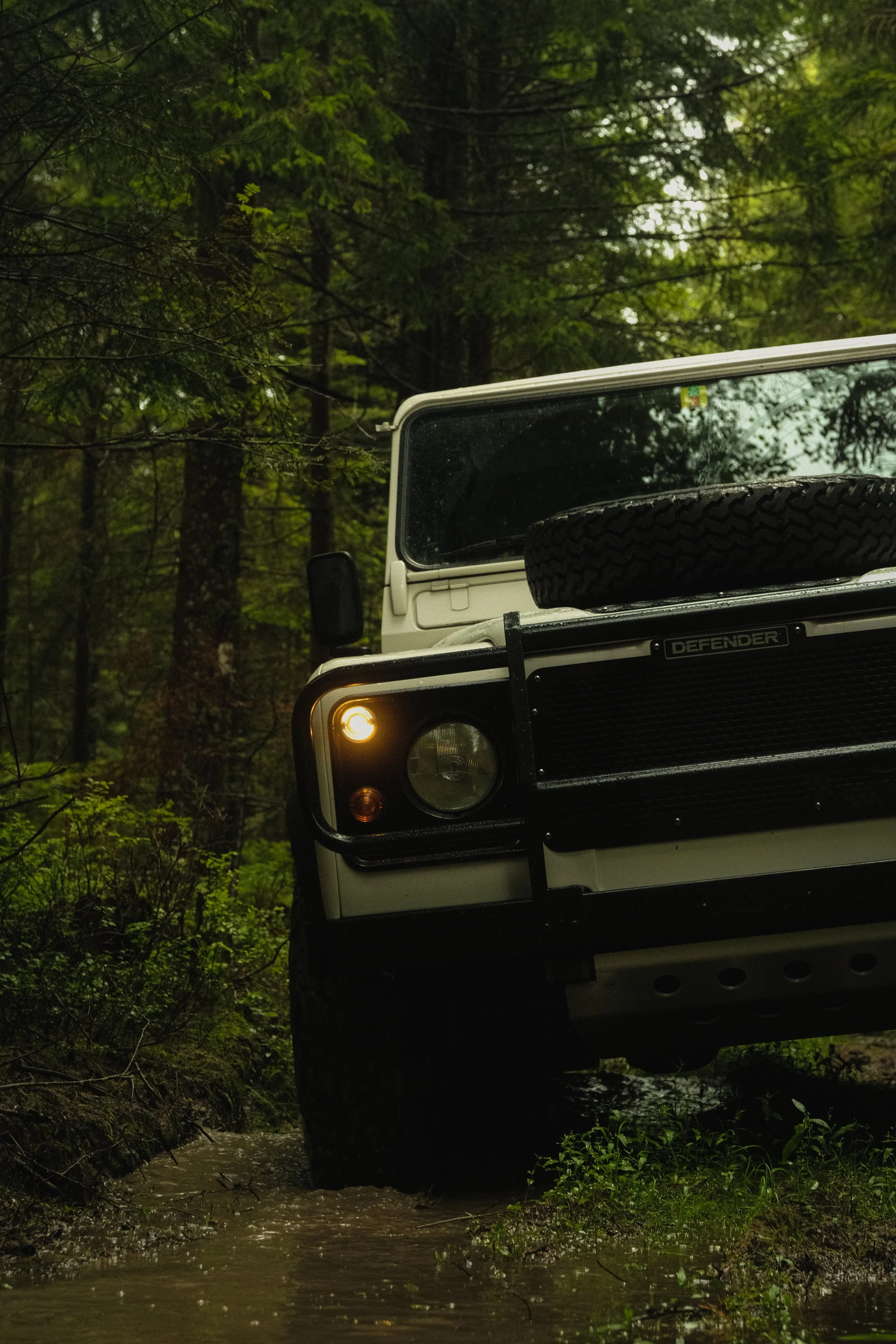 Ein weißer Geländewagen, der in einem Wald auf schlammigem Boden steht, mit einem voll aufgestellten Reserverad auf der Motorhaube.