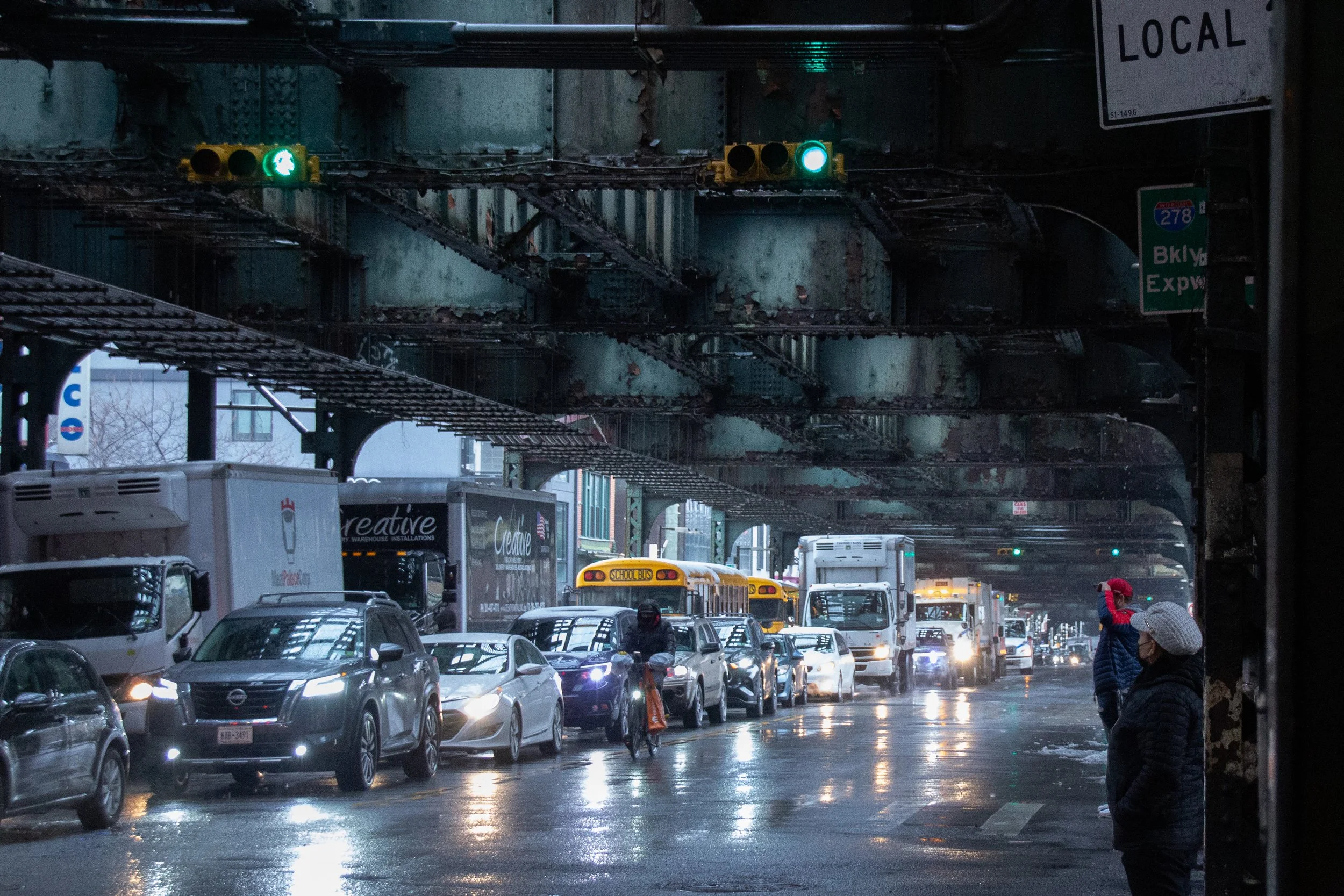 Stau auf einer nassen, regnerischen Stadtstraße unter einer alten Eisenbahnbrücke, mit Autos, Bussen und Fußgängern, einige tragen Winterkleidung